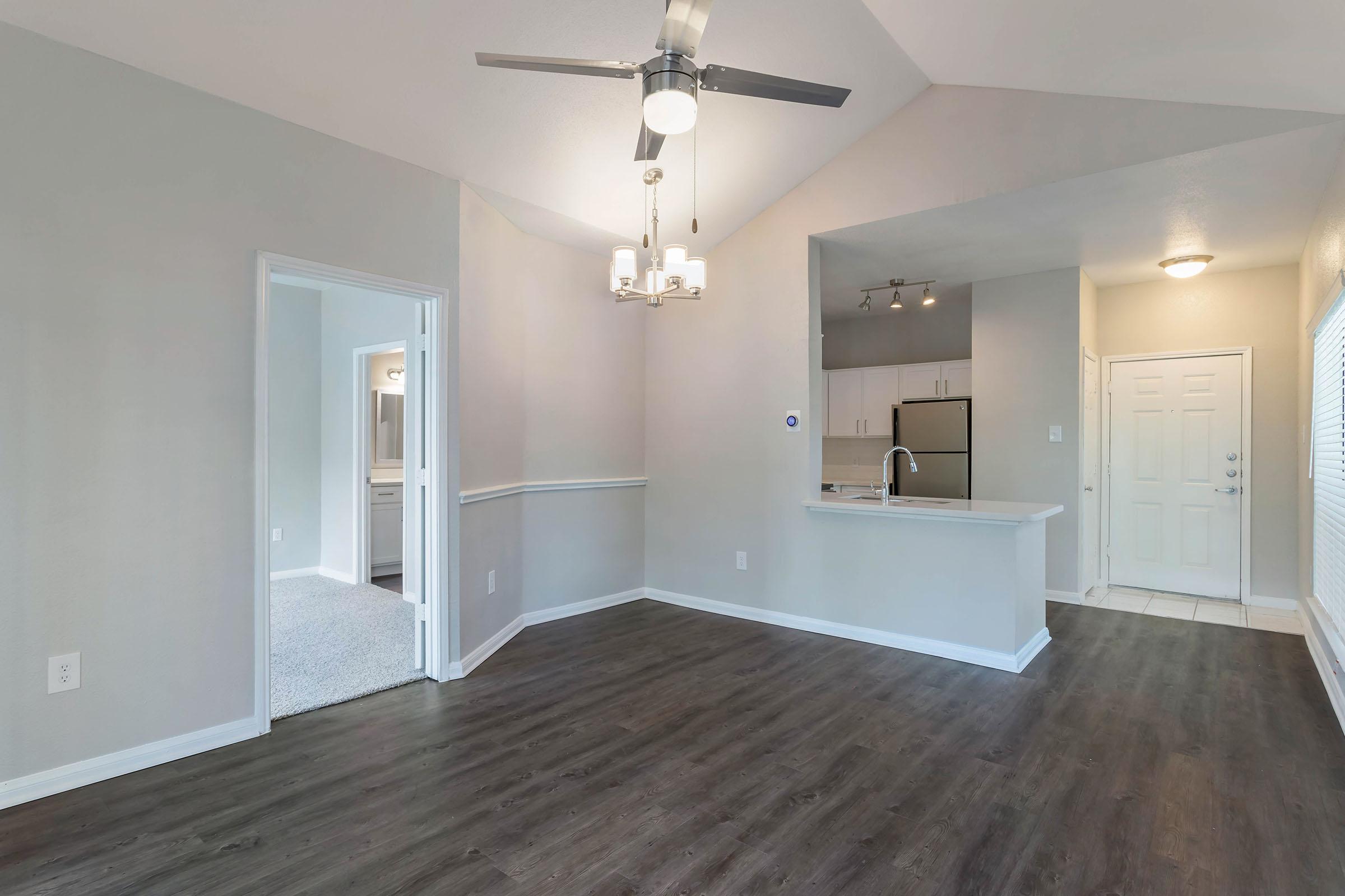 Modern interior of a living space featuring a ceiling fan, neutral-colored walls, and laminate flooring. A doorway leads to another room, and the kitchen area is visible with stainless steel appliances. The entrance door is on the right, and there is a small chandelier above the dining area.