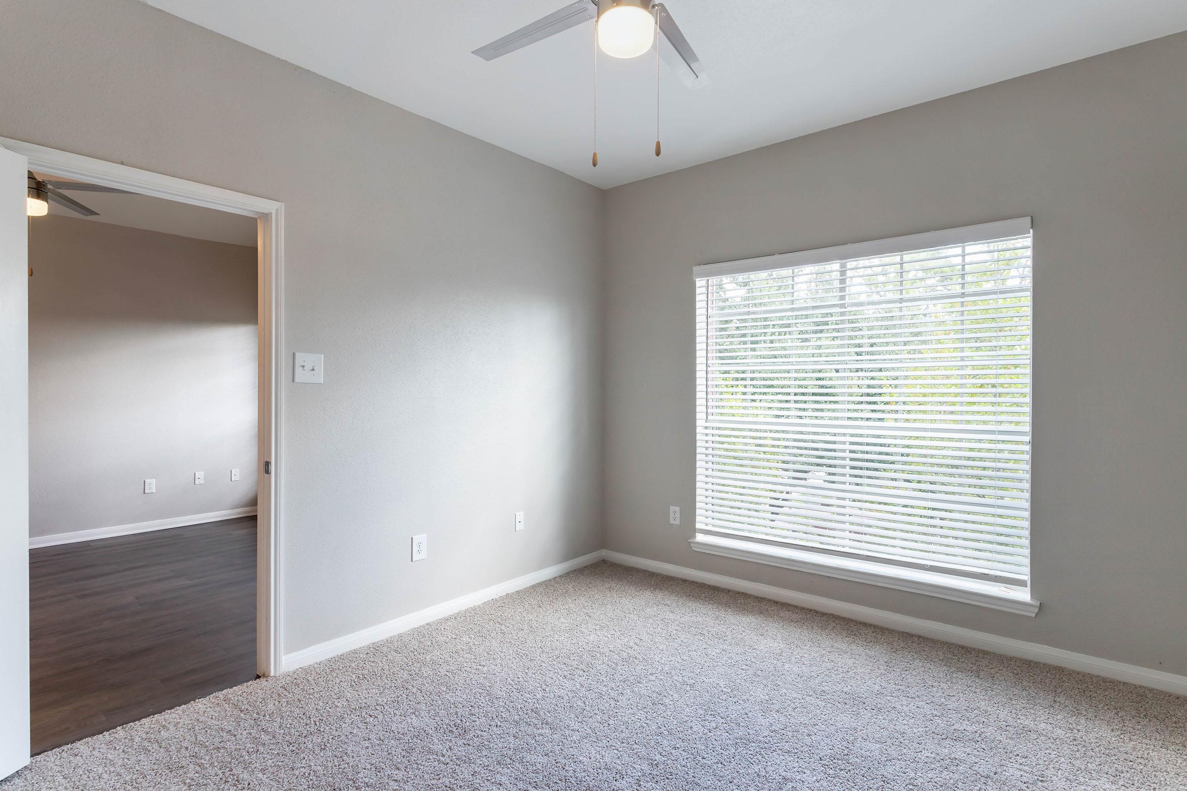 A light gray bedroom with a ceiling fan and a window featuring blinds. The carpet is beige, and there is an open doorway leading to another room. The walls are plain and the room is well-lit, creating a spacious feel.