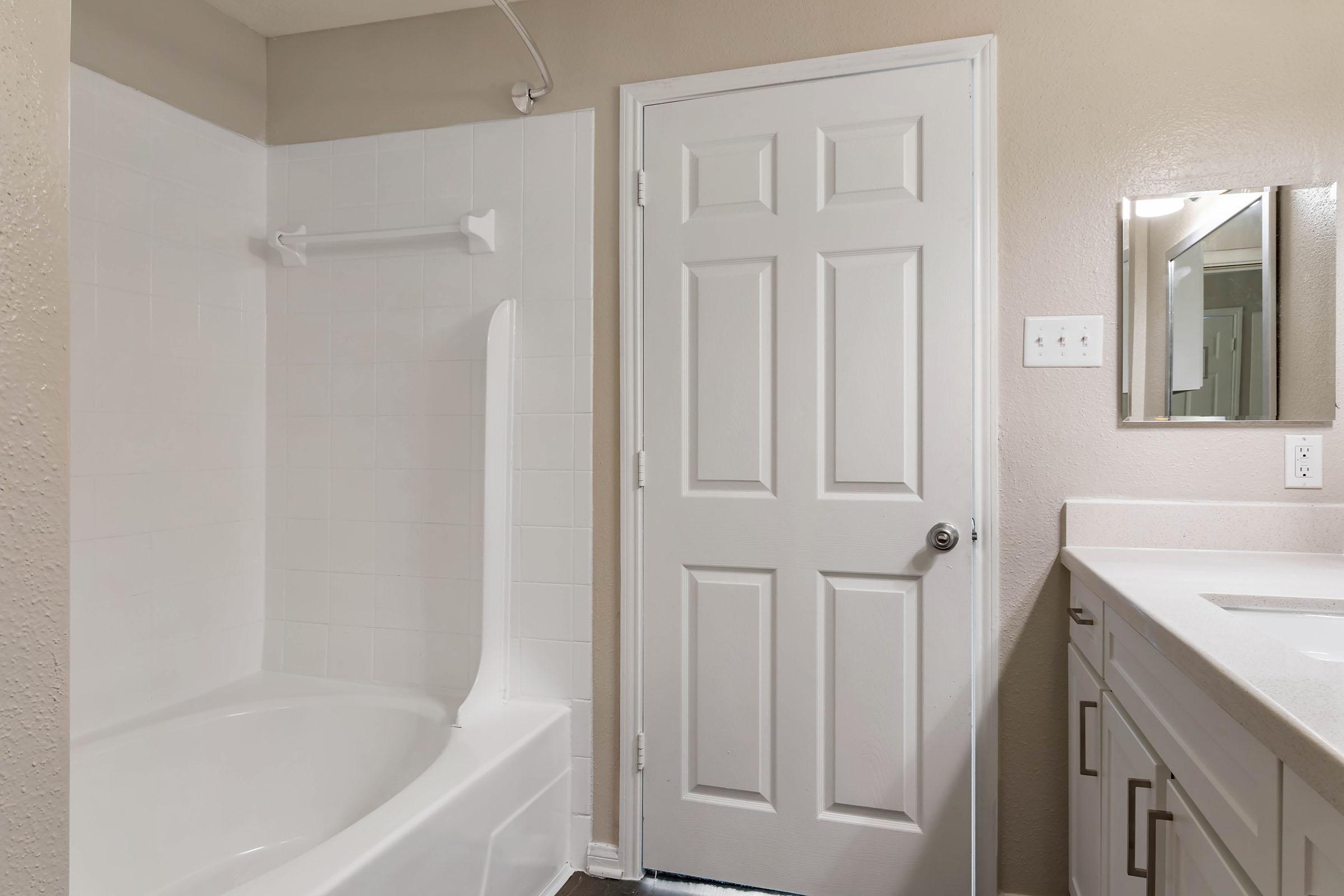 A clean and modern bathroom featuring a bathtub with a white tile surround, a closed white door, and a vanity with a mirror. The walls are painted in a light beige color, and the flooring appears to be a dark wood or laminate. Natural light brightens the space, creating a welcoming atmosphere.