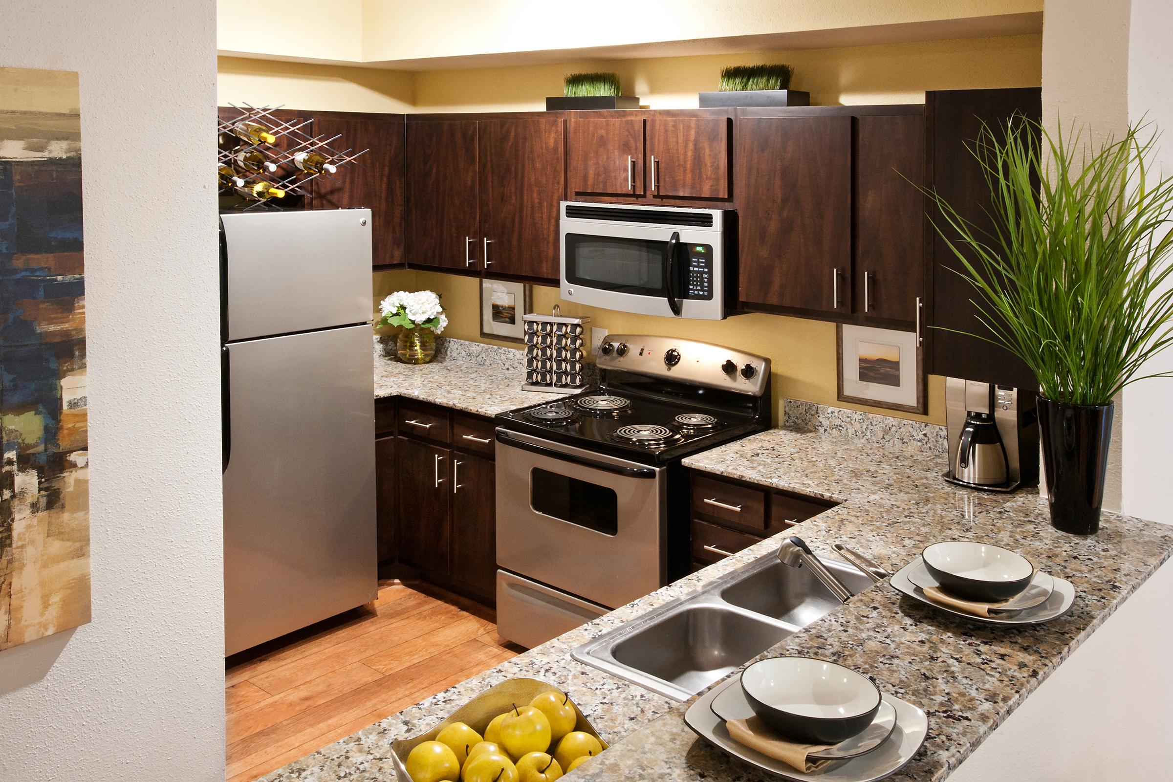 Modern kitchen featuring dark wooden cabinetry, stainless steel appliances, and a granite countertop. A sink is visible along with bowls and decorative items. There are green plants and a bowl of lemons on the countertop, and artwork on the walls. The space is well-lit with a warm ambiance.