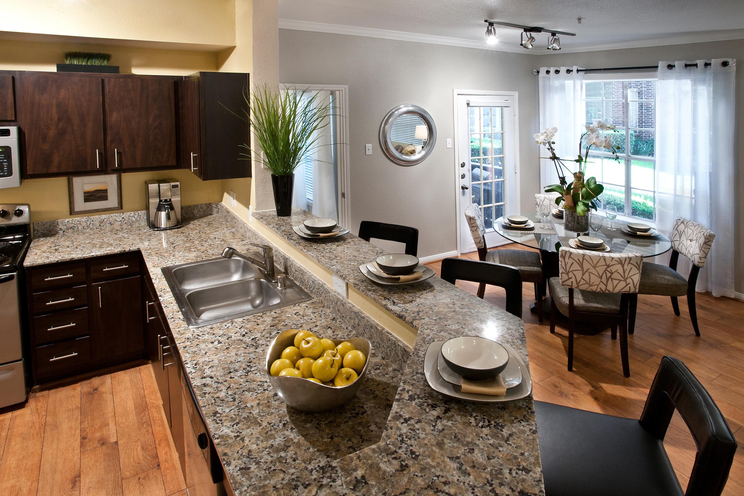 A modern kitchen with a granite countertop featuring a bowl of lemons. Stainless steel appliances and dark wood cabinets are visible. Adjacent is a dining area with a round table set with plates and chairs, surrounded by large windows letting in natural light.