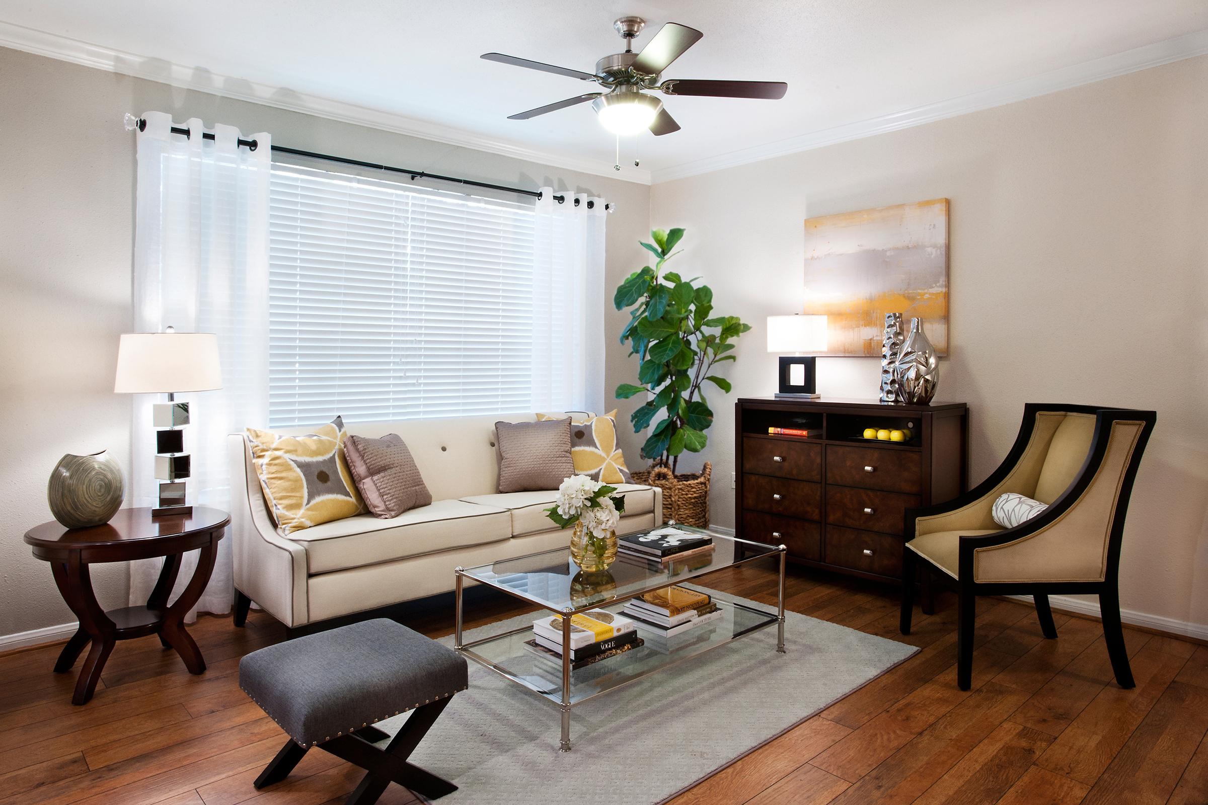 Bright and airy living room with a cream-colored sofa adorned with decorative pillows, a glass coffee table, and a stylish armchair. A potted plant adds a touch of greenery, while a modern ceiling fan and lamps provide warm lighting. A wooden dresser and artwork complete the cozy atmosphere.