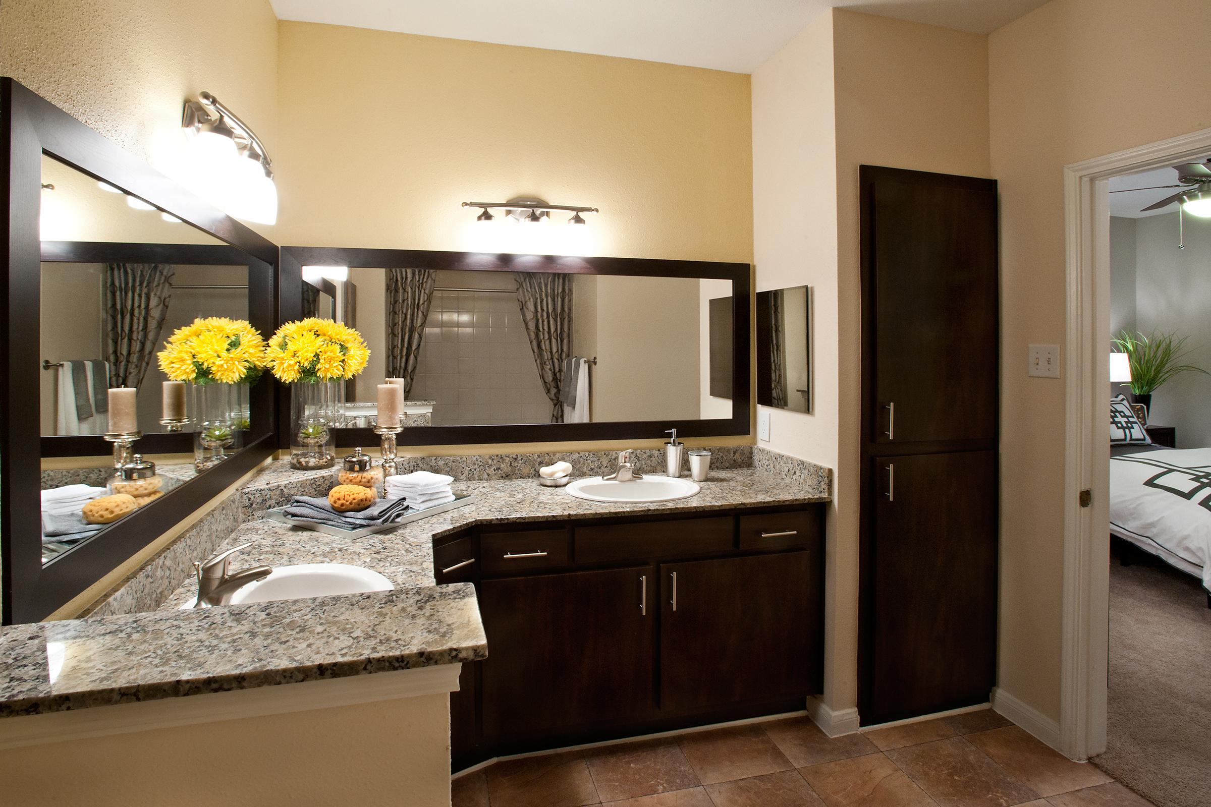 A modern bathroom featuring a granite countertop with a sink, decorative yellow flowers in a vase, candles, and towels. Mirrors are mounted on the wall above the countertop, and there is a spacious cabinet nearby. The adjoining room is partially visible, showcasing a bed with neutral bedding.
