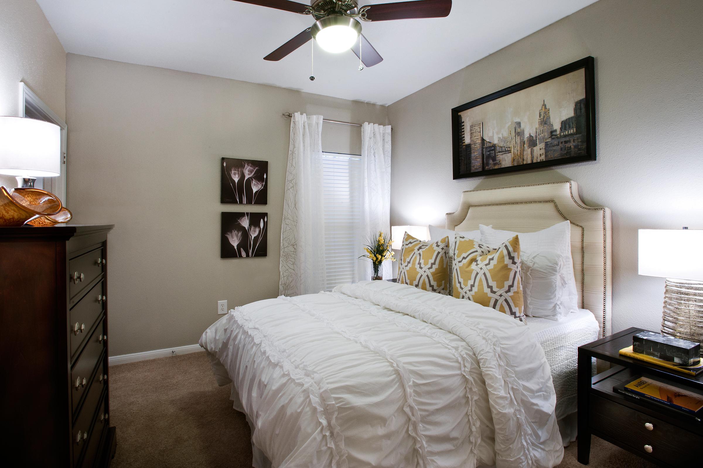 A cozy bedroom featuring a queen-sized bed with white bedding and decorative pillows. There are two framed floral artworks on the wall, a wooden dresser, and a nightstand with a lamp. Natural light filters through a window with sheer curtains, and a ceiling fan provides ventilation.