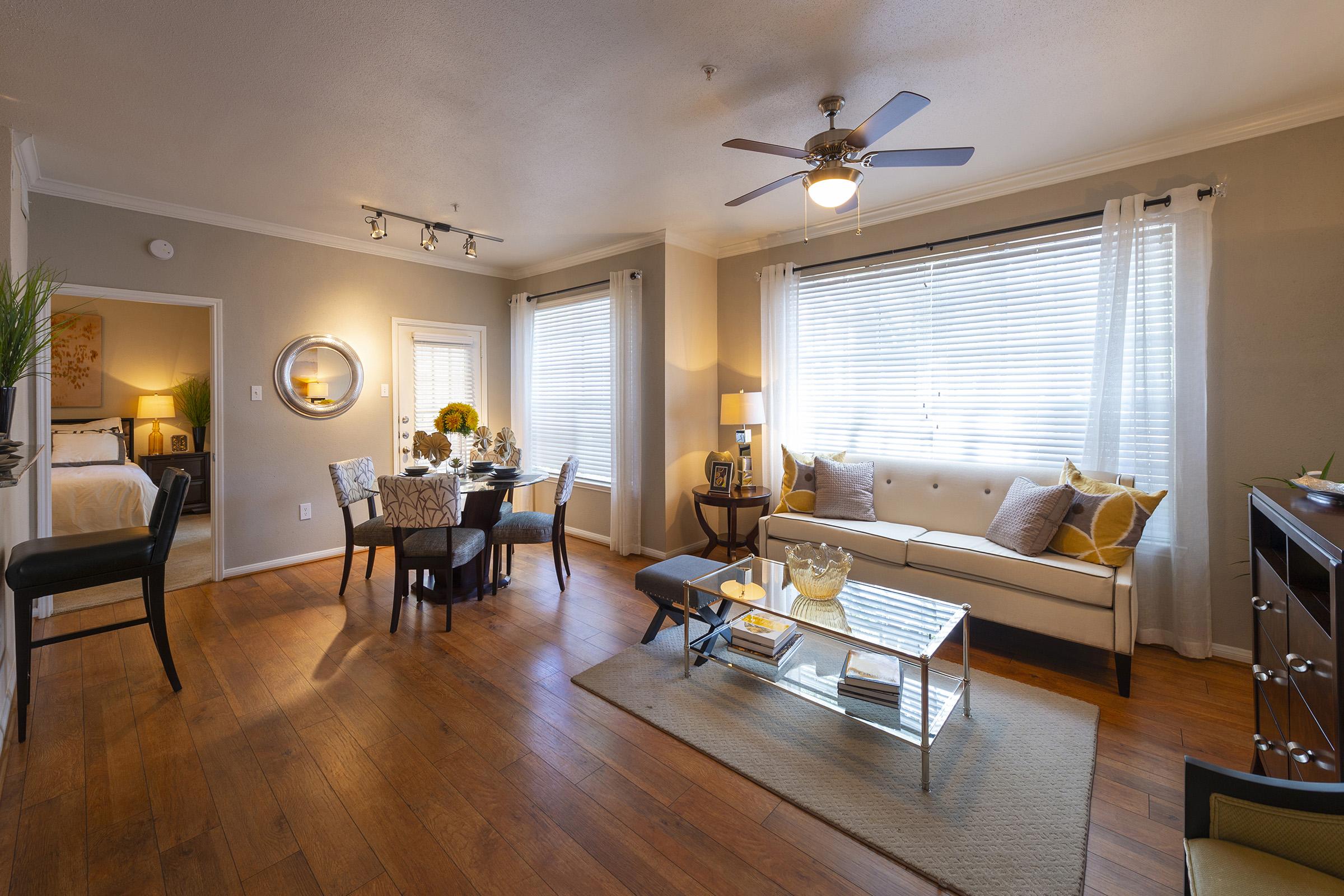 A modern living space featuring a comfortable sofa, a glass coffee table, and a dining area with a round table set for four. Natural light streams through large windows with sheer curtains, illuminating the wooden floor. Decorative items like cushions and plants add warmth to the inviting atmosphere.