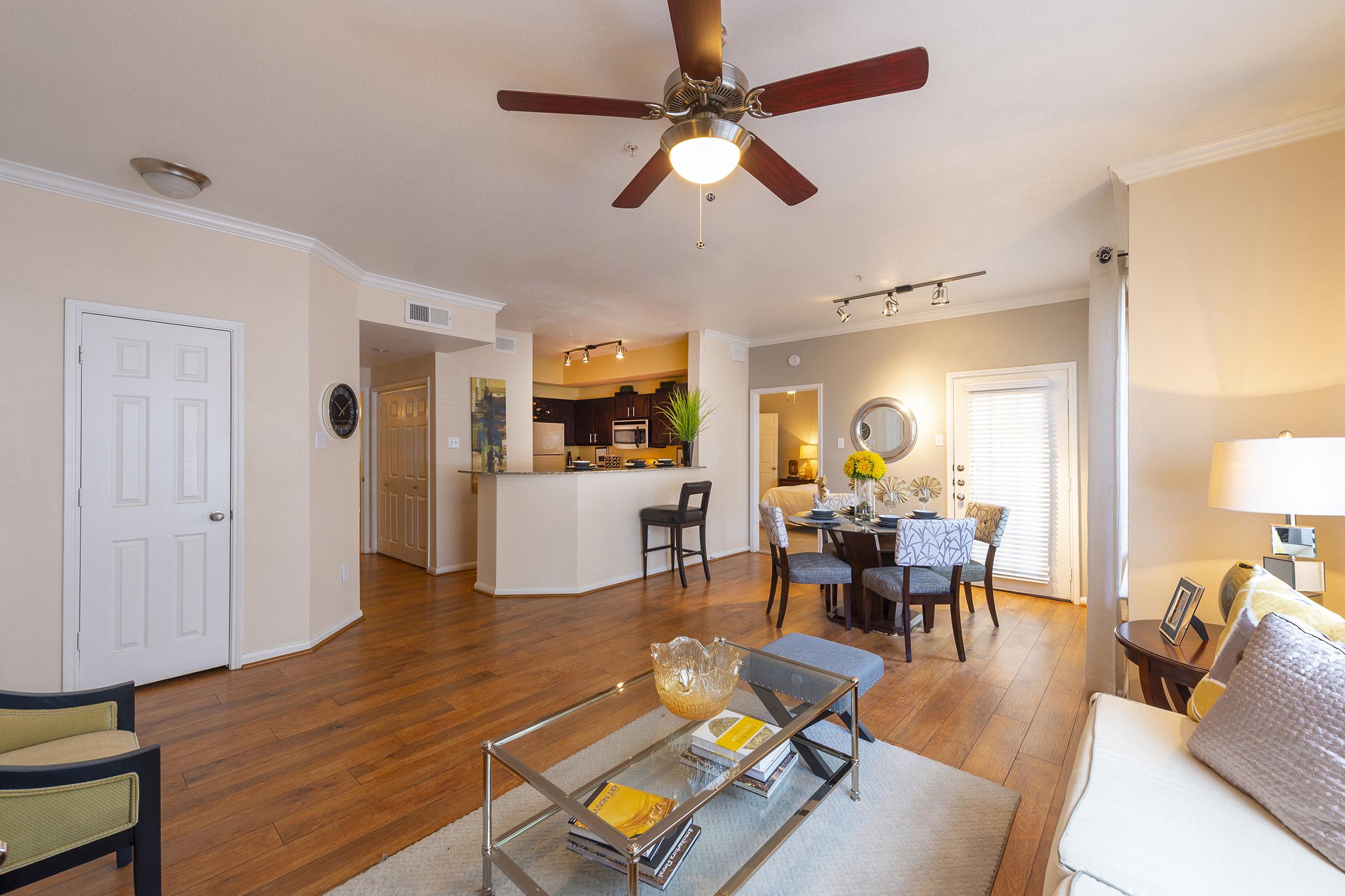 Interior view of a modern living room featuring hardwood floors, a ceiling fan, and a cozy seating area. A glass coffee table sits in the center, surrounded by stylish chairs. The dining area with dark wood furniture is visible in the background, along with a kitchen that includes contemporary cabinetry. Natural light enters through sliding doors.