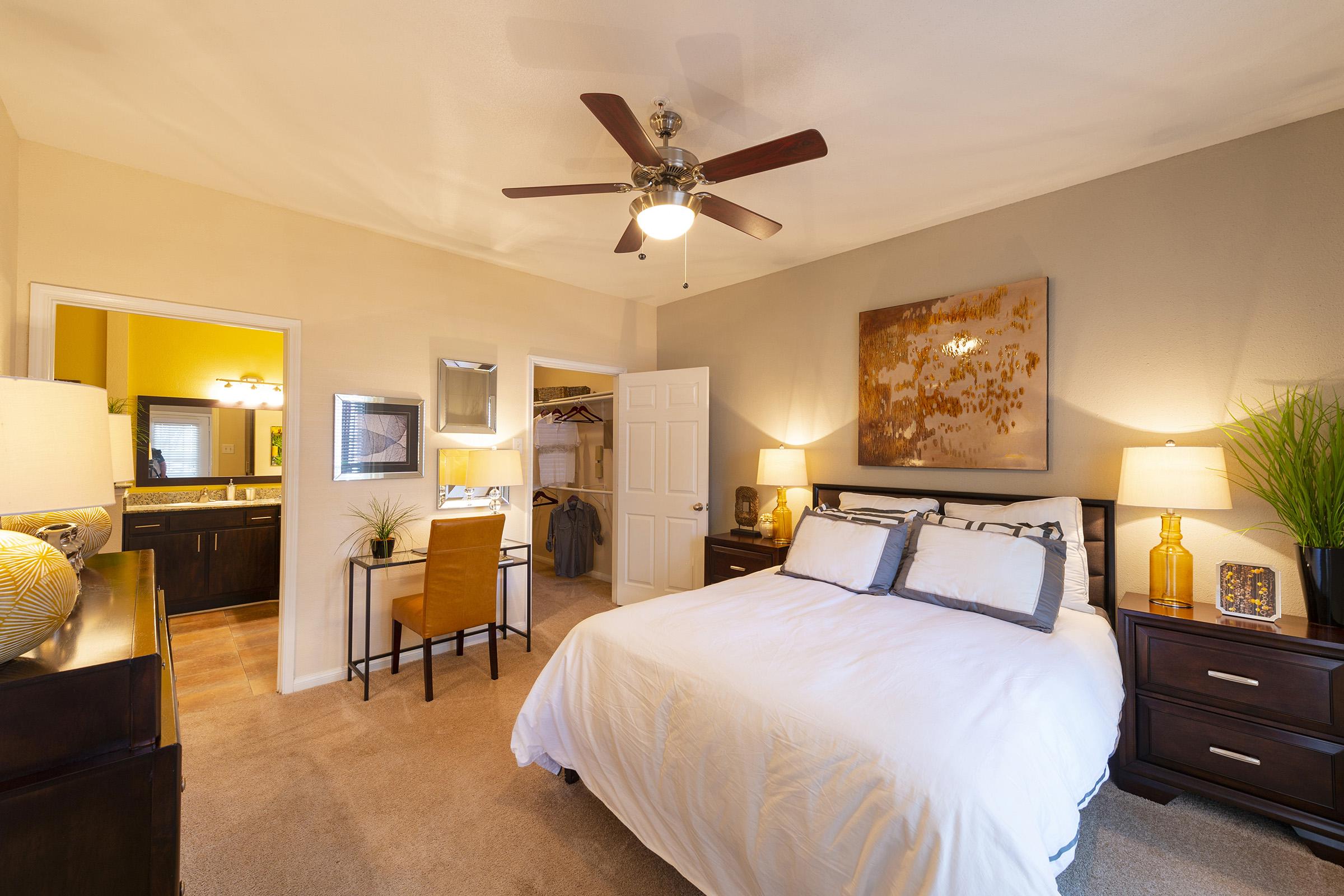 A well-decorated bedroom featuring a large bed with white linens, two bedside tables with lamps, a ceiling fan, and a framed artwork on the wall. A mirror and a desk with a chair are visible, along with an open closet in the background, contributing to a cozy and inviting atmosphere.