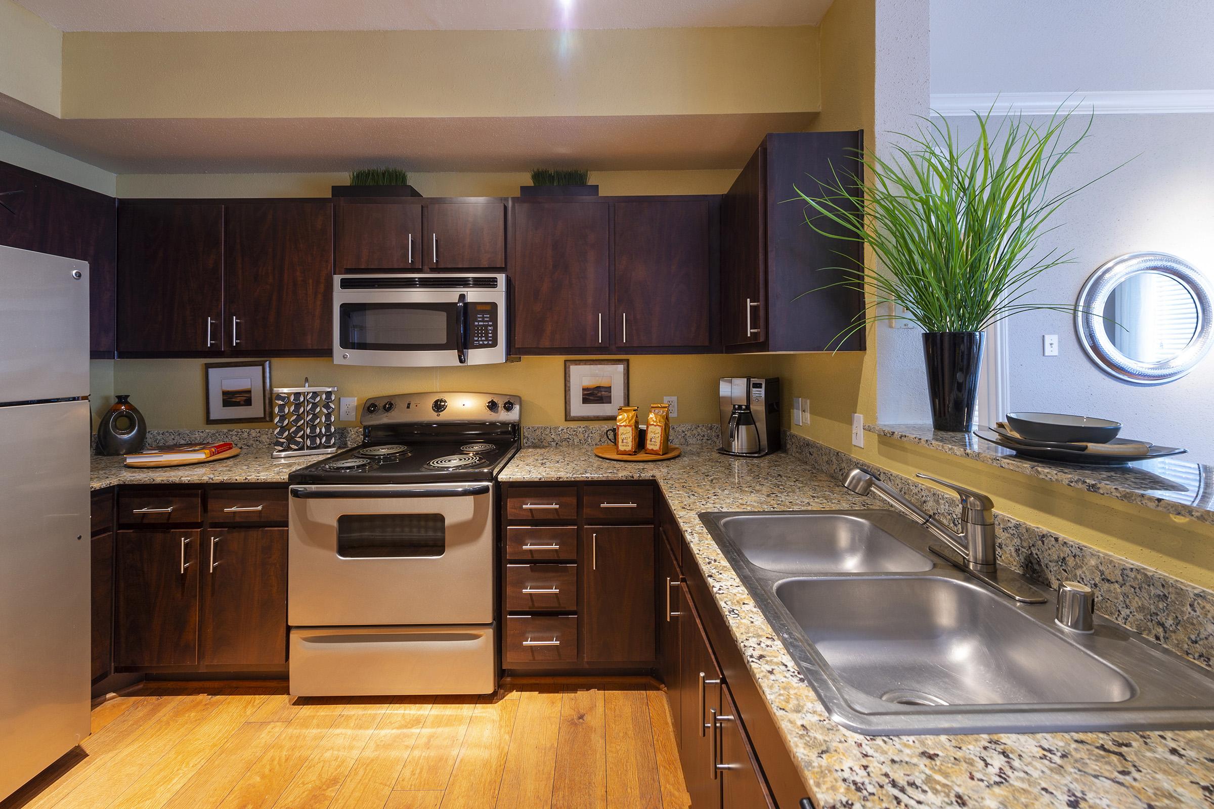 Modern kitchen featuring dark wooden cabinets, stainless steel appliances, and a granite countertop. Includes a double sink, a stove with an overhead microwave, and decorative elements like a potted plant and circular mirror. The warm wooden floor complements the contemporary design.