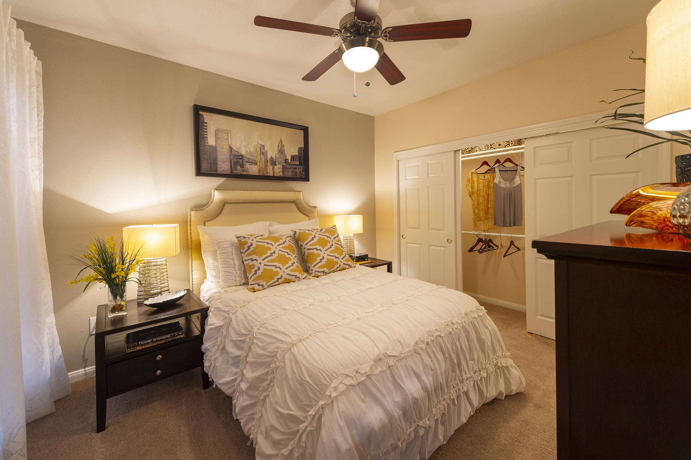 Cozy bedroom featuring a neatly made bed with a white ruffled duvet and yellow accent pillows. Two bedside tables with lamps are placed on either side, and a fan is mounted on the ceiling. A closet with white doors is seen in the background, displaying hangers with clothes. A plant adds a touch of greenery to the space.