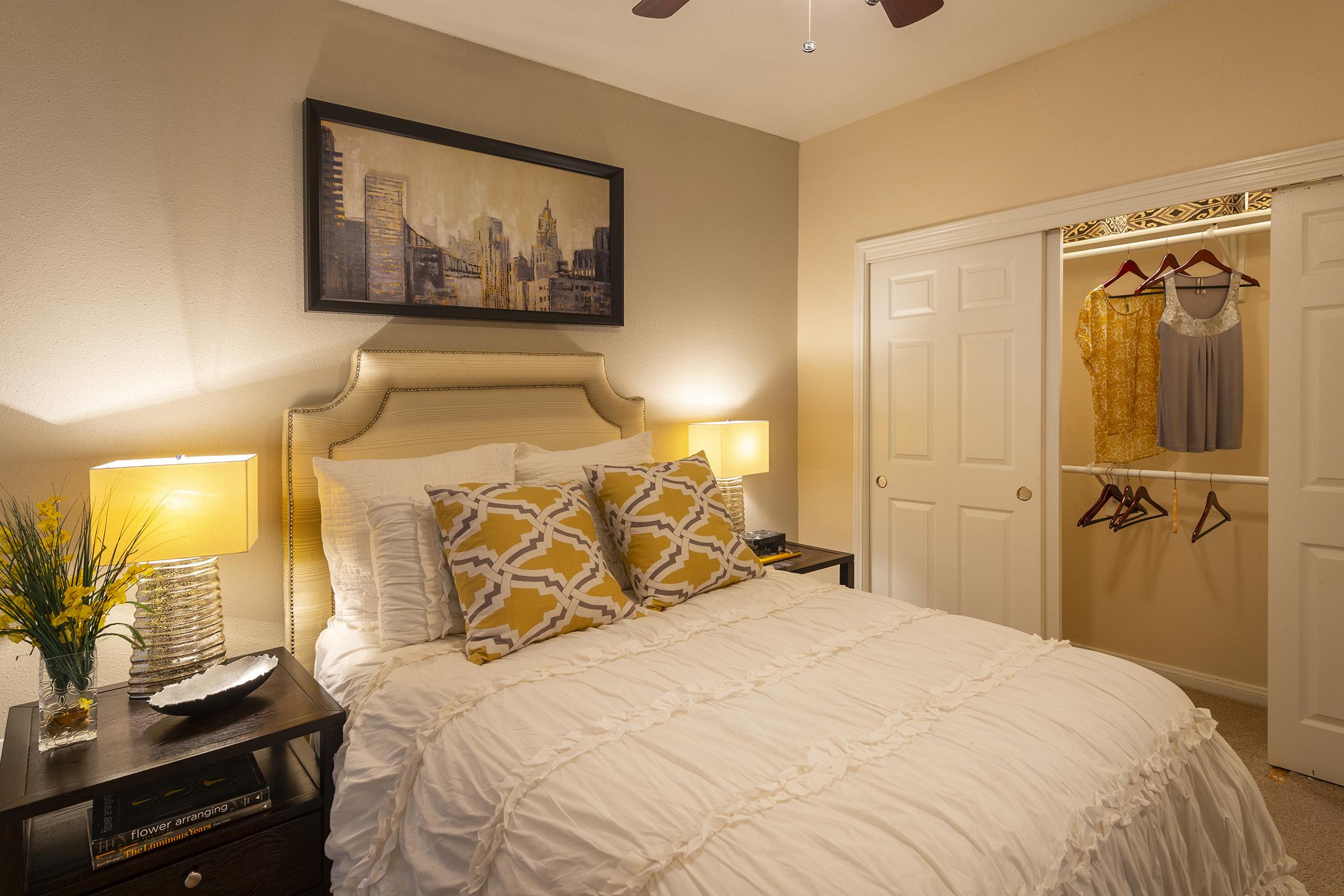 Cozy bedroom featuring a neatly made bed with white ruffled bedding and yellow accent pillows. There are two bedside lamps illuminating the space, and a framed cityscape painting on the wall. A closet door is slightly open, displaying hanging clothes and a top shelf. A small vase with greenery is on the nightstand.