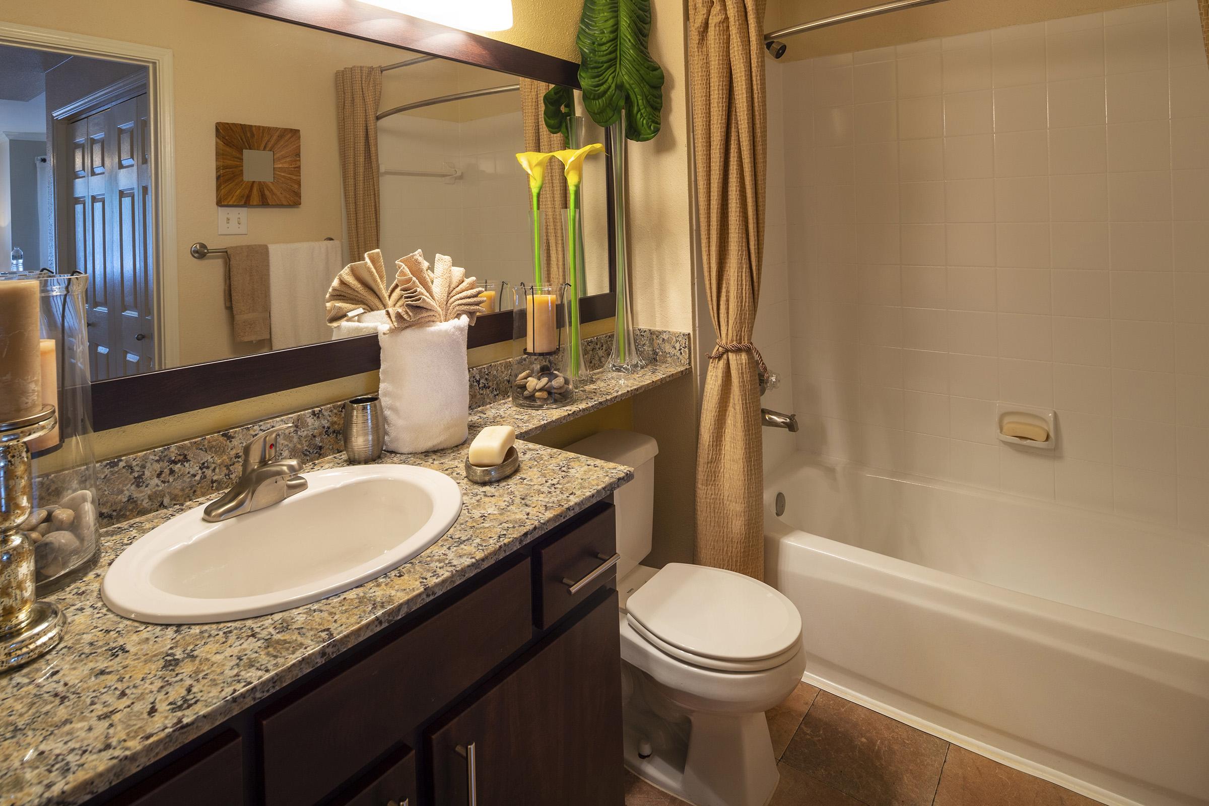 A well-lit bathroom featuring a granite countertop with a sink, a modern toilet, and a bathtub with a shower curtain. Decorative elements include a vase with yellow flowers, a shell display, and soft towels hanging on the wall. The overall ambiance is clean and inviting.