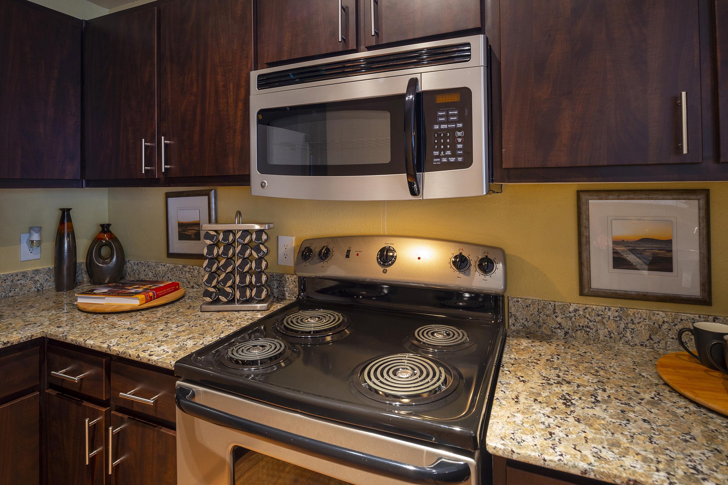 A modern kitchen featuring dark wood cabinetry and a granite countertop. The image shows a stainless steel microwave above a black stovetop with visible burners. A wine rack and decorative items are placed on the counter, and framed pictures are mounted on the wall, adding a cozy touch to the space.