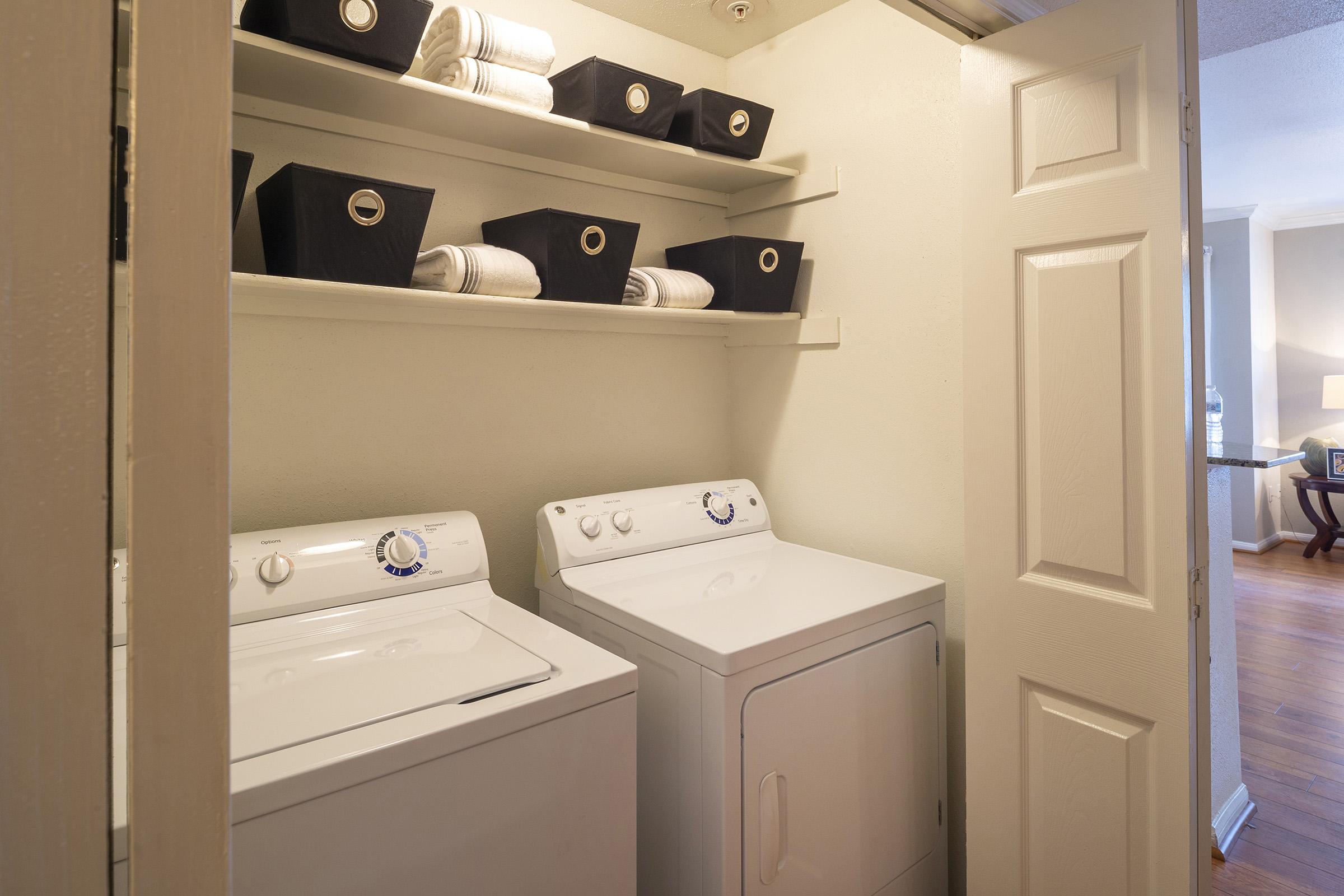 A laundry room featuring a stacked washer and dryer with white cabinetry. Above, there are shelves holding neatly rolled towels and black storage bins. The room has a light-colored wall, and the doorway opens to a living space with hardwood floors visible in the background.