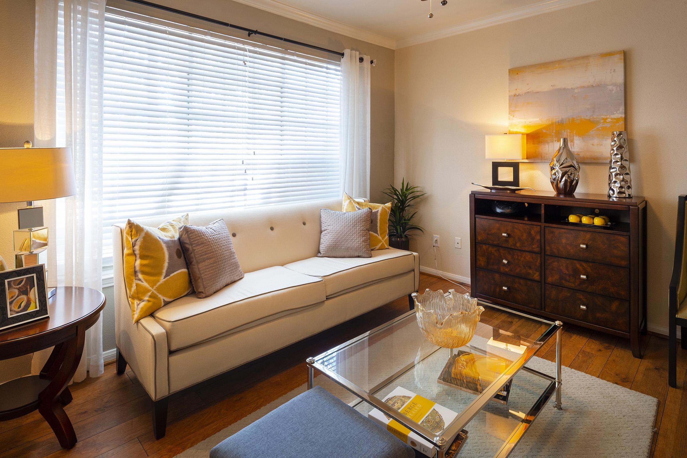A cozy living room featuring a beige couch with decorative pillows, a glass coffee table, and a wooden cabinet. Natural light flows in through a window with white blinds, complemented by a plant and a table lamp, creating a warm and inviting atmosphere.