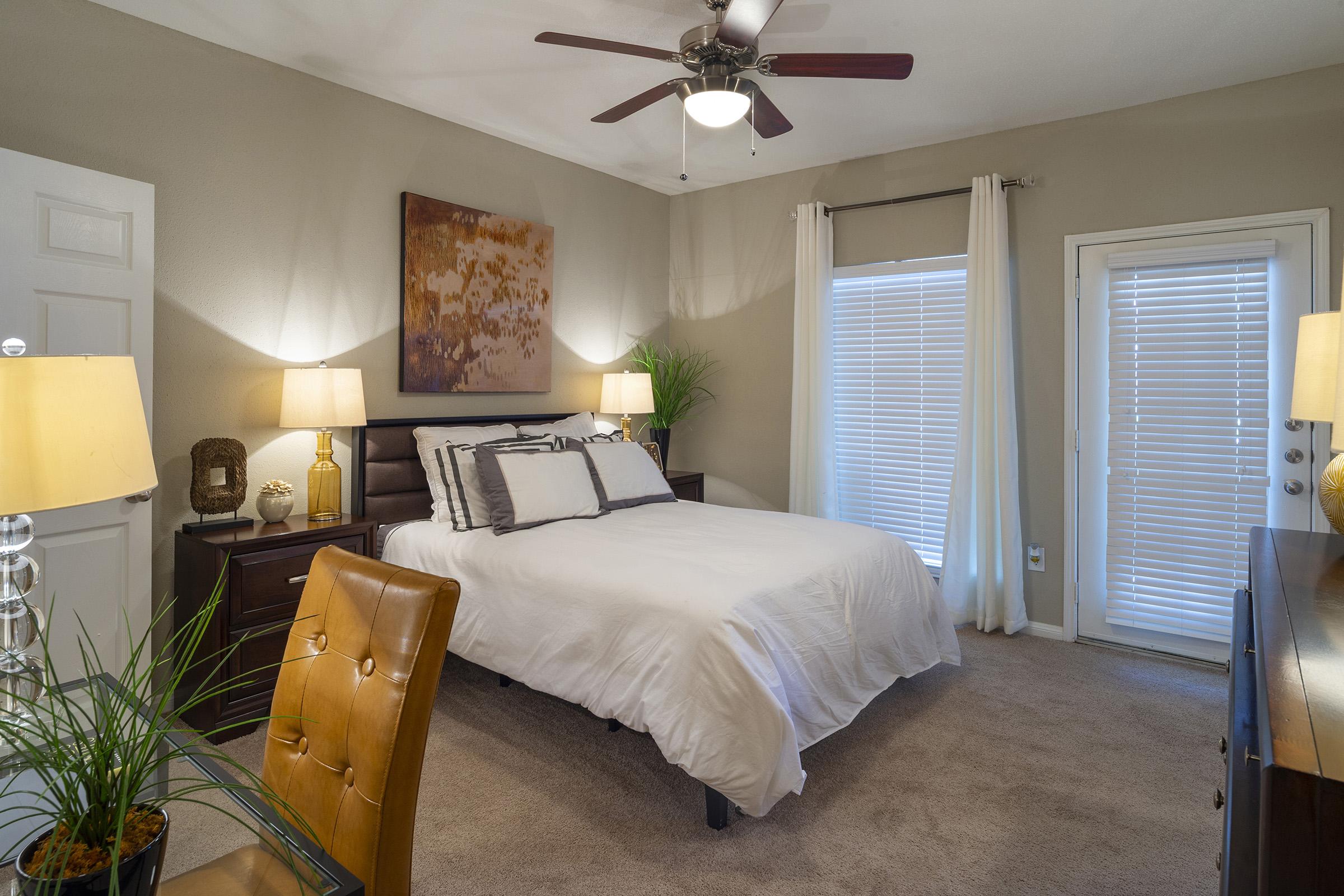 A cozy bedroom featuring a queen-sized bed with white bedding, bedside lamps, and a modern ceiling fan. There are two windows with blinds, a decorative painting on the wall, and a small desk with a brown chair. The room has neutral wall colors and a carpeted floor, creating a warm and inviting atmosphere.