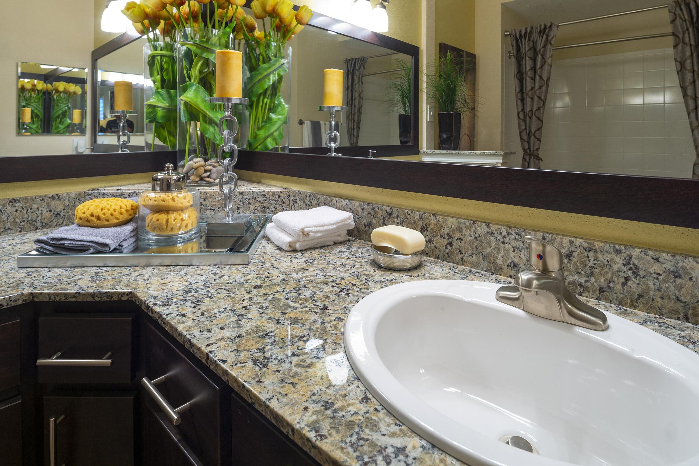 A modern bathroom countertop featuring a sink, granite surface, decorative yellow flowers, candles, a glass jar with decorative stones, and neatly folded white towels. The mirrors above reflect the warm lighting and plants, creating a welcoming atmosphere.