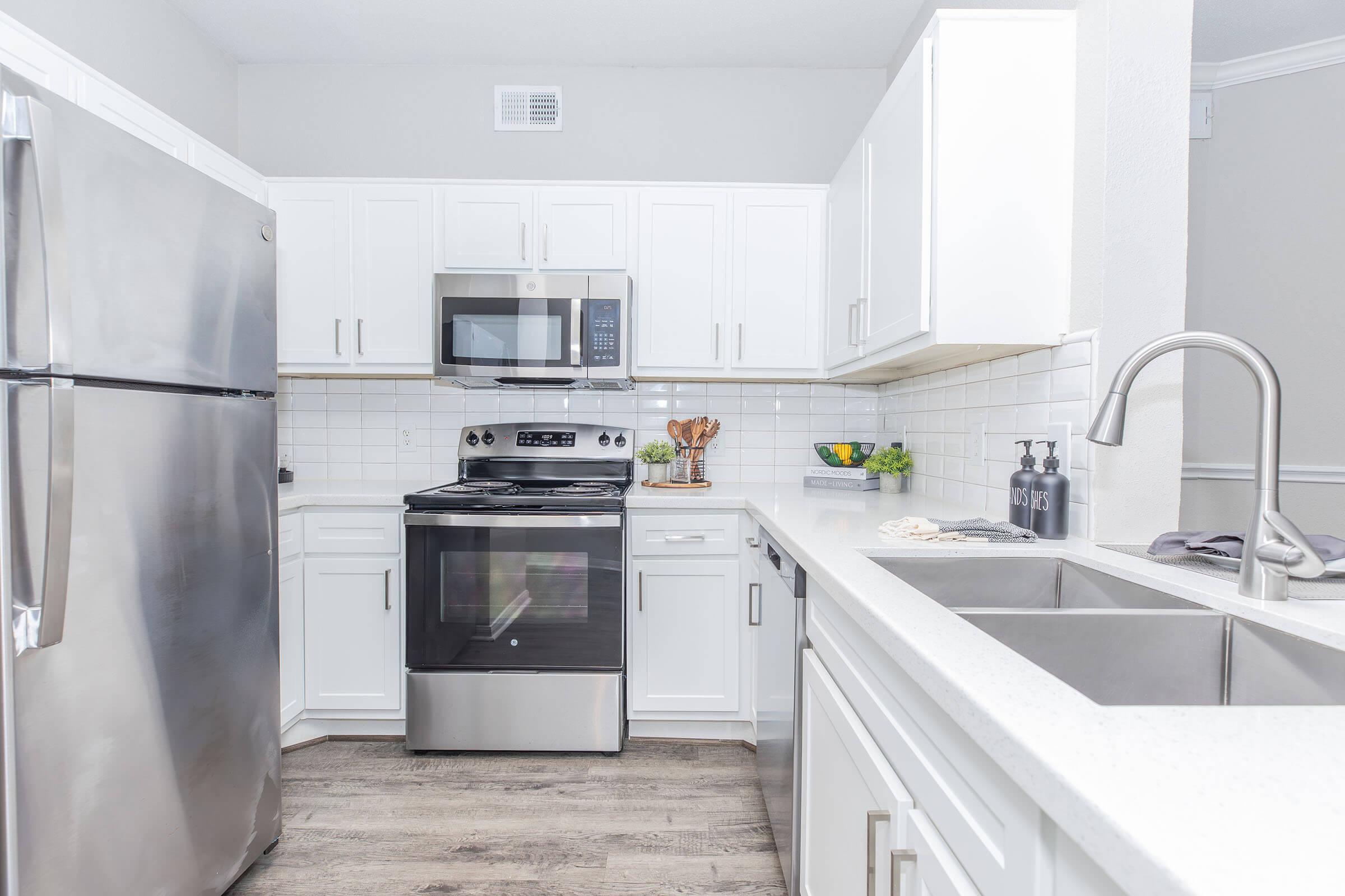 Modern kitchen with sleek stainless steel appliances, including a refrigerator, oven, and microwave. White cabinets and a light-colored countertop create a bright atmosphere. A double sink is positioned near the window, and decorative items like bowls of fruit add a touch of color. The flooring is light wood, enhancing the contemporary feel.