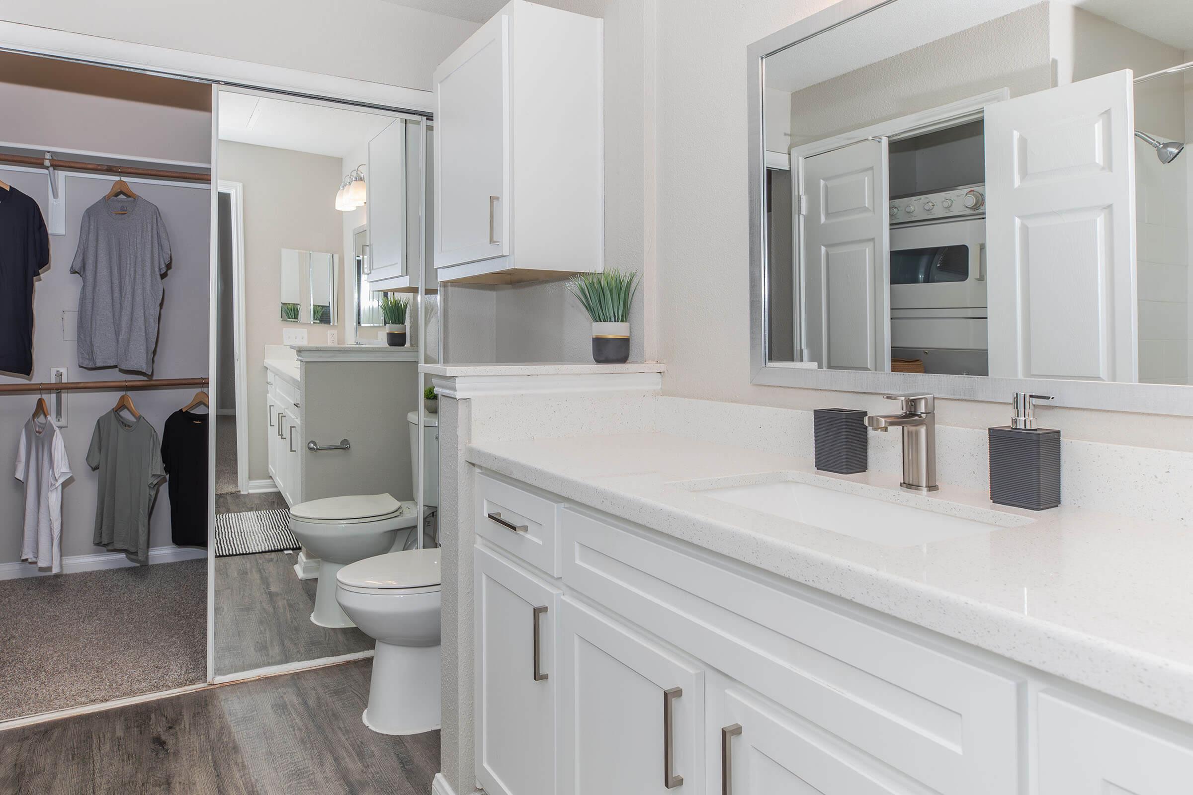 A modern bathroom featuring a double sink vanity with a white countertop, a large mirror, and stylish fixtures. To the side, there's a closet area with hanging clothes. The space is well-lit with neutral colors and contemporary design, including a toilet and a small decorative plant.
