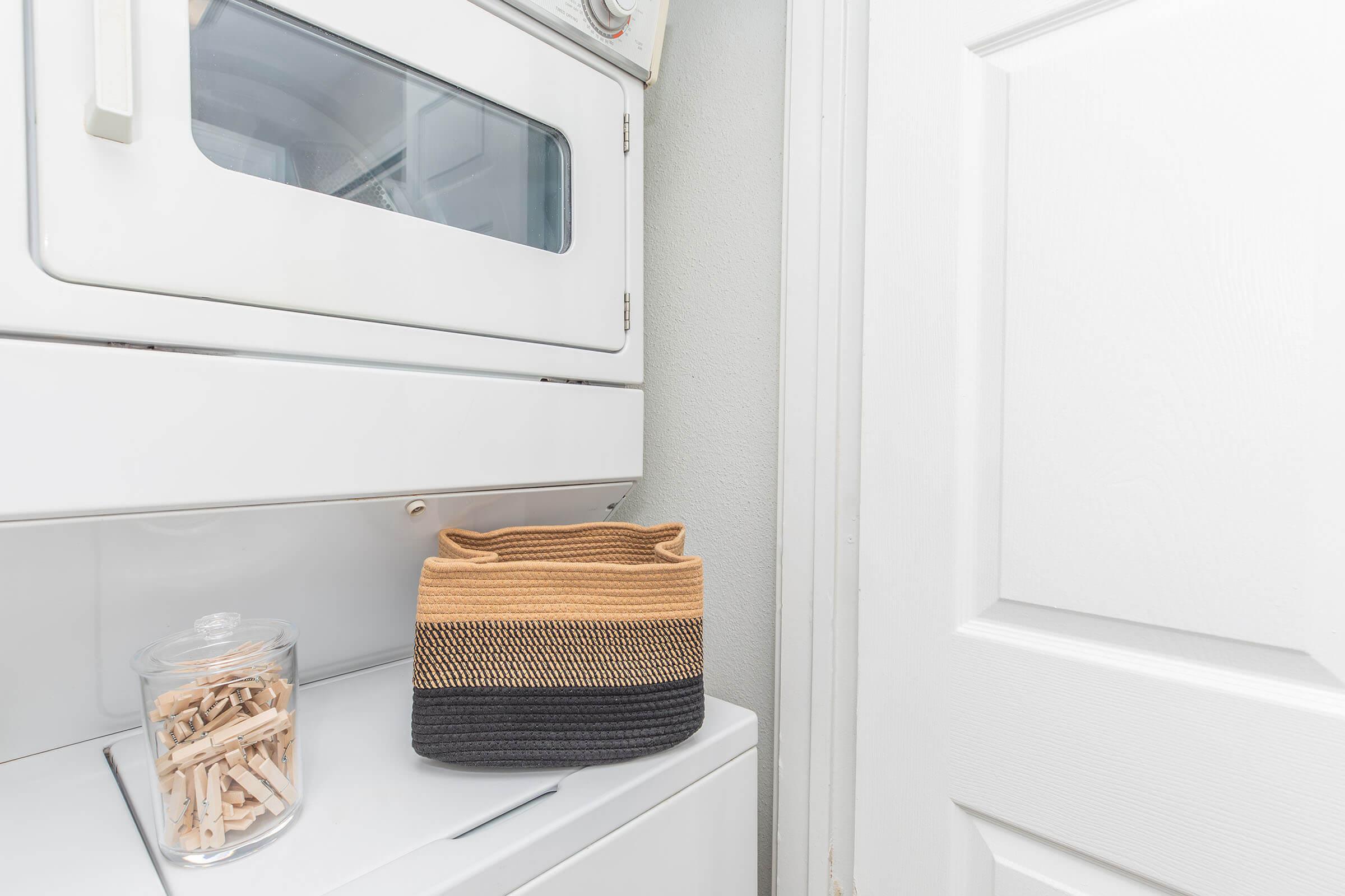 A laundry area featuring stacked washer and dryer units, a white door, and a basket made of woven material in black and tan colors. Nearby, a glass jar filled with clothespins is placed on the surface. The wall is painted in a light color, creating a clean and organized space.