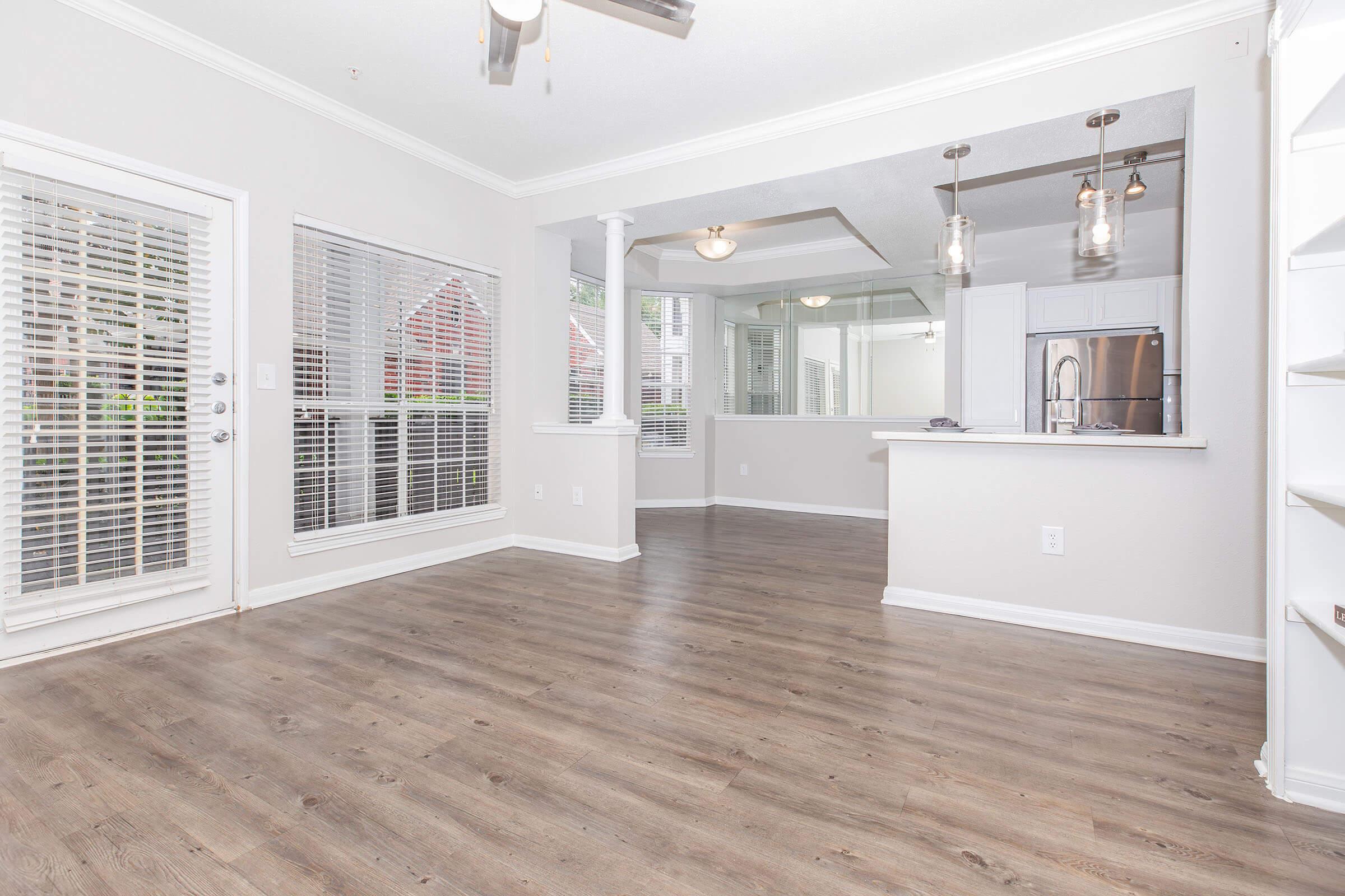 Bright and open living space featuring large windows with white shutters, light-colored walls, and hardwood flooring. The room transitions into a kitchen area with modern pendant lighting and an open layout, creating a warm and inviting atmosphere.