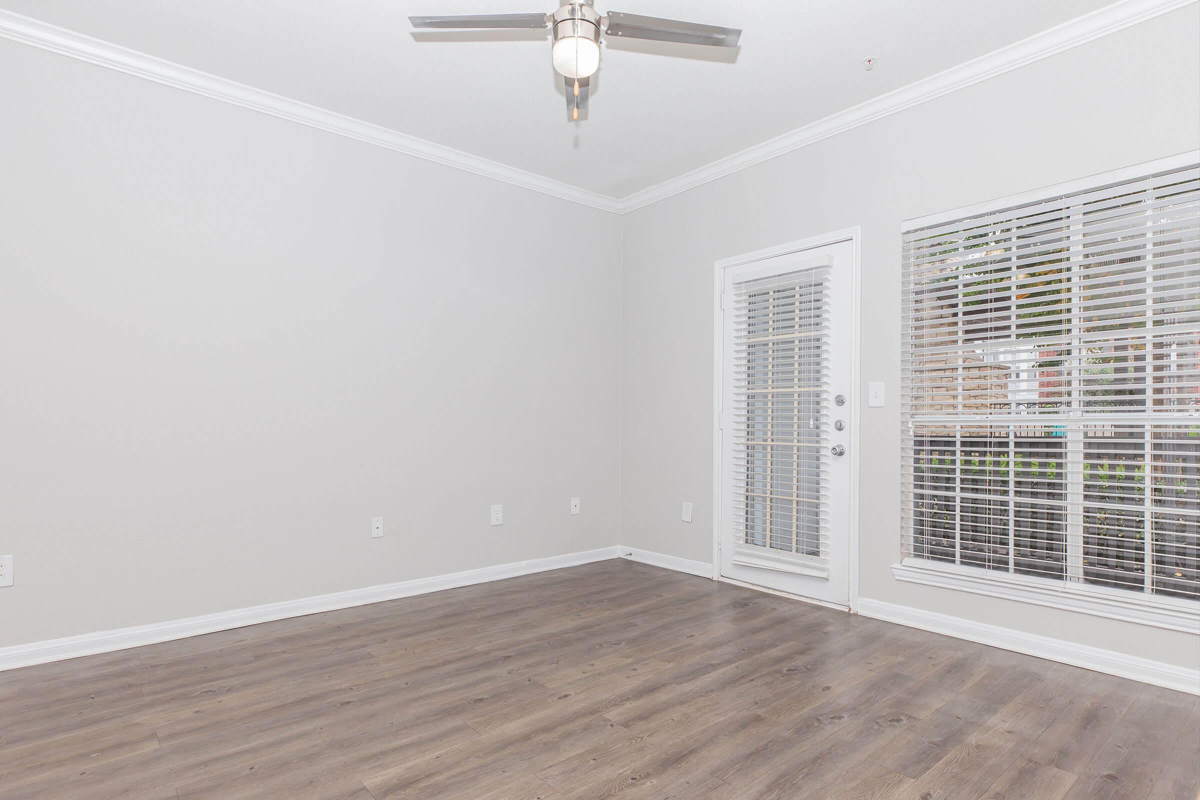 Empty room with light gray walls and hardwood flooring, featuring a ceiling fan and a large window with blinds. A door leads to an outdoor area, providing natural light. The space is open and uncluttered, ideal for various interior design possibilities.