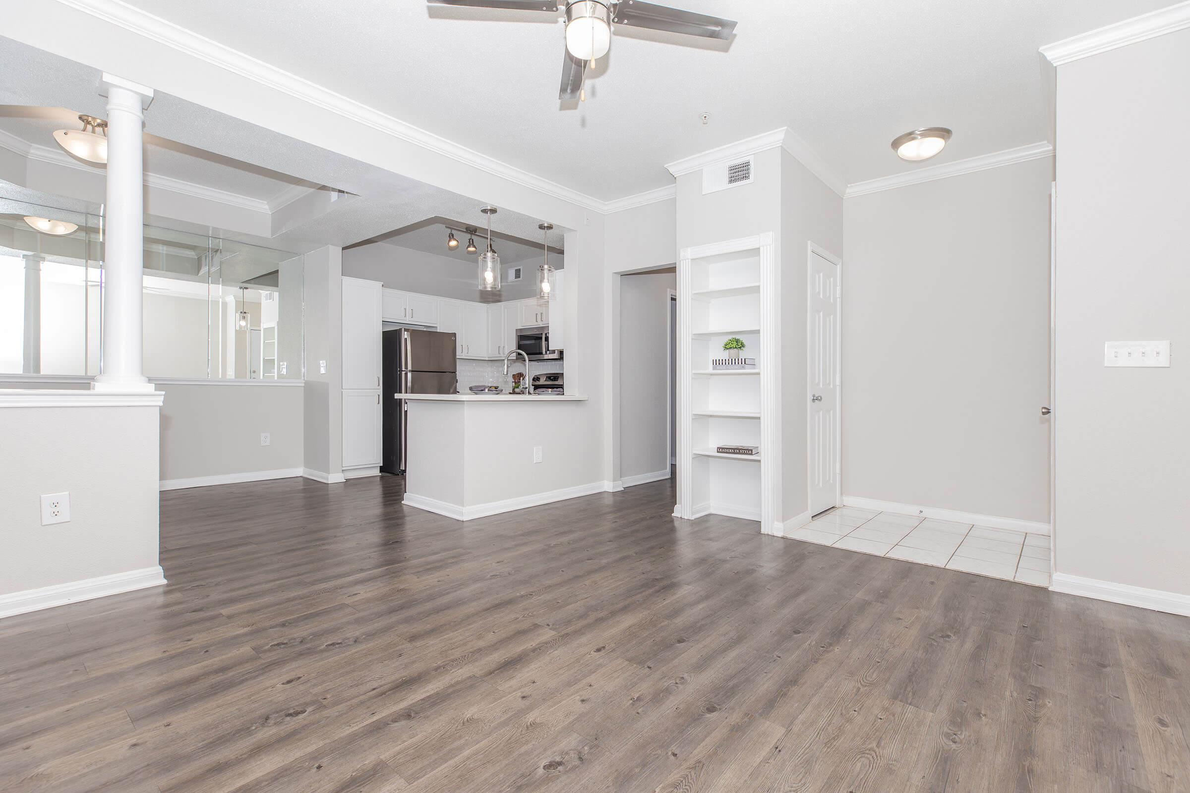 Spacious living area with wooden flooring, featuring a ceiling fan. The open layout includes a modern kitchen with stainless steel appliances, a white cabinet, and a doorway leading to a tiled entryway. Natural light fills the room through large windows.