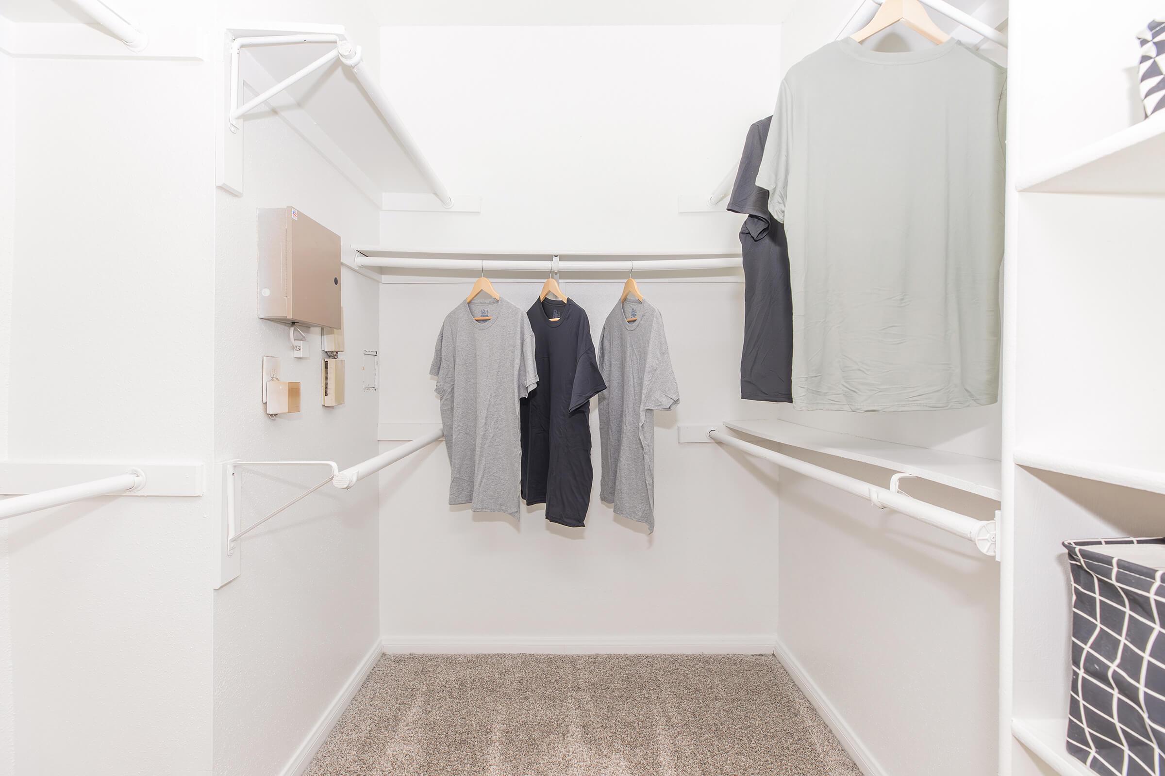 A neatly organized closet featuring several hanging t-shirts in neutral colors. The closet has white walls and shelving, with a light-colored carpet on the floor. There are no other items visible, creating a clean and minimalist appearance.