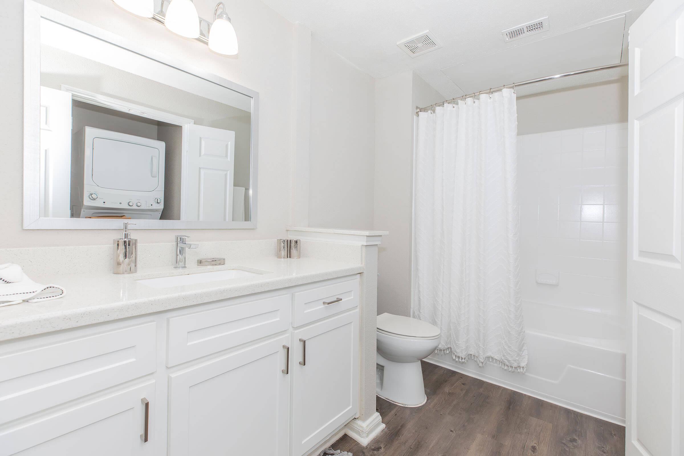 A clean, modern bathroom featuring a white vanity with a sink, large mirror, and elegant fixtures. There’s a shower with a white curtain and a toilet. Light wood-style flooring enhances the bright atmosphere, and a washing machine is discreetly placed in a nook near the shower.