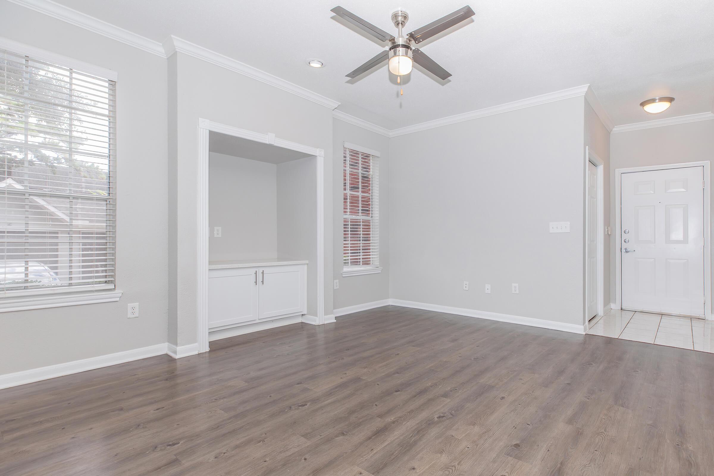 A spacious, empty living room featuring light gray walls, a ceiling fan, and large windows with blinds. There is a built-in cabinet on one side and a tile entryway visible, leading to the front door. The flooring is a rich wood-style laminate, creating a modern and inviting atmosphere.