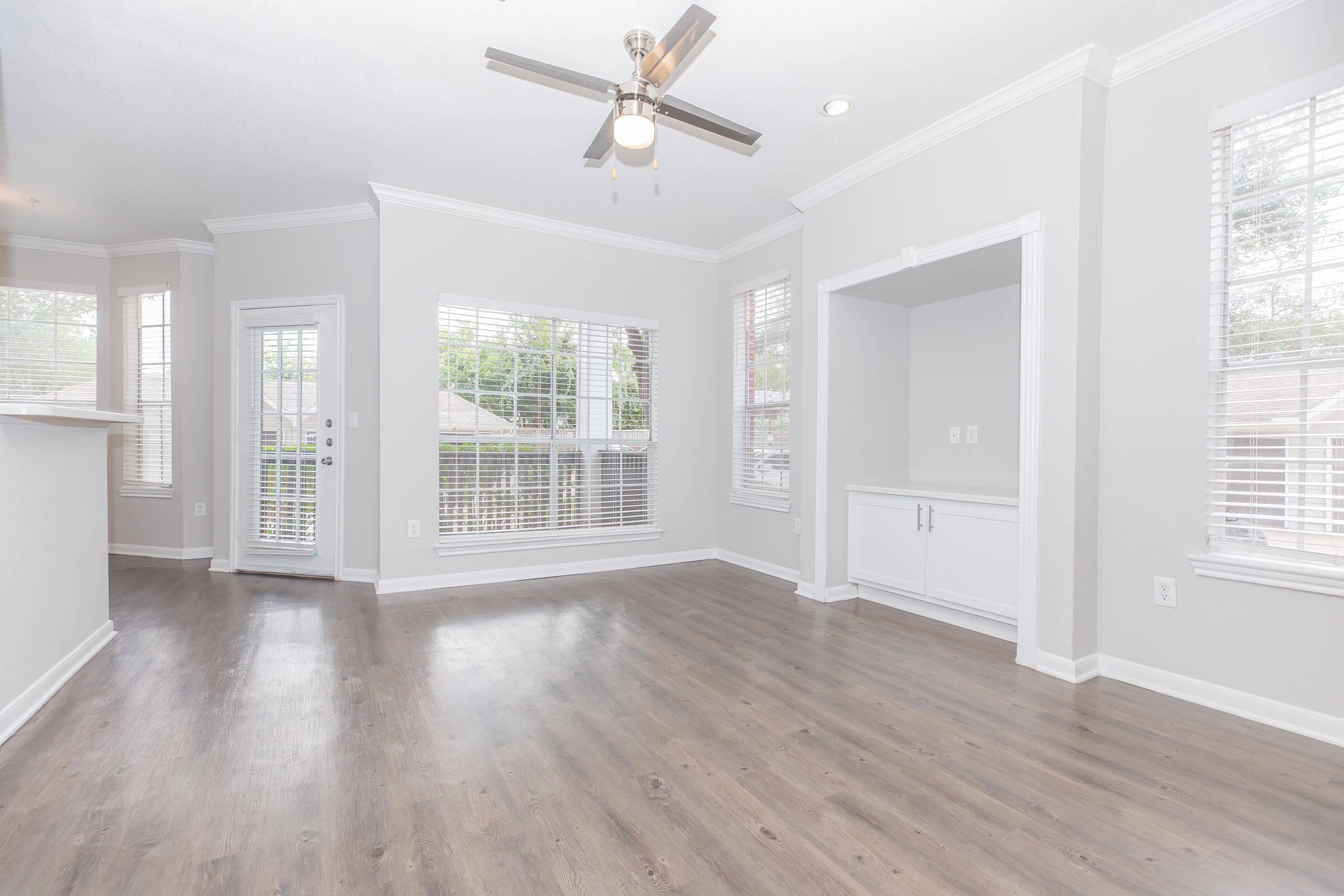 A modern living room featuring light gray walls, large windows with blinds, and a ceiling fan. The floor is made of dark wood laminate. There's a built-in cabinet on one wall and a door leading out, providing ample natural light and a spacious feel.