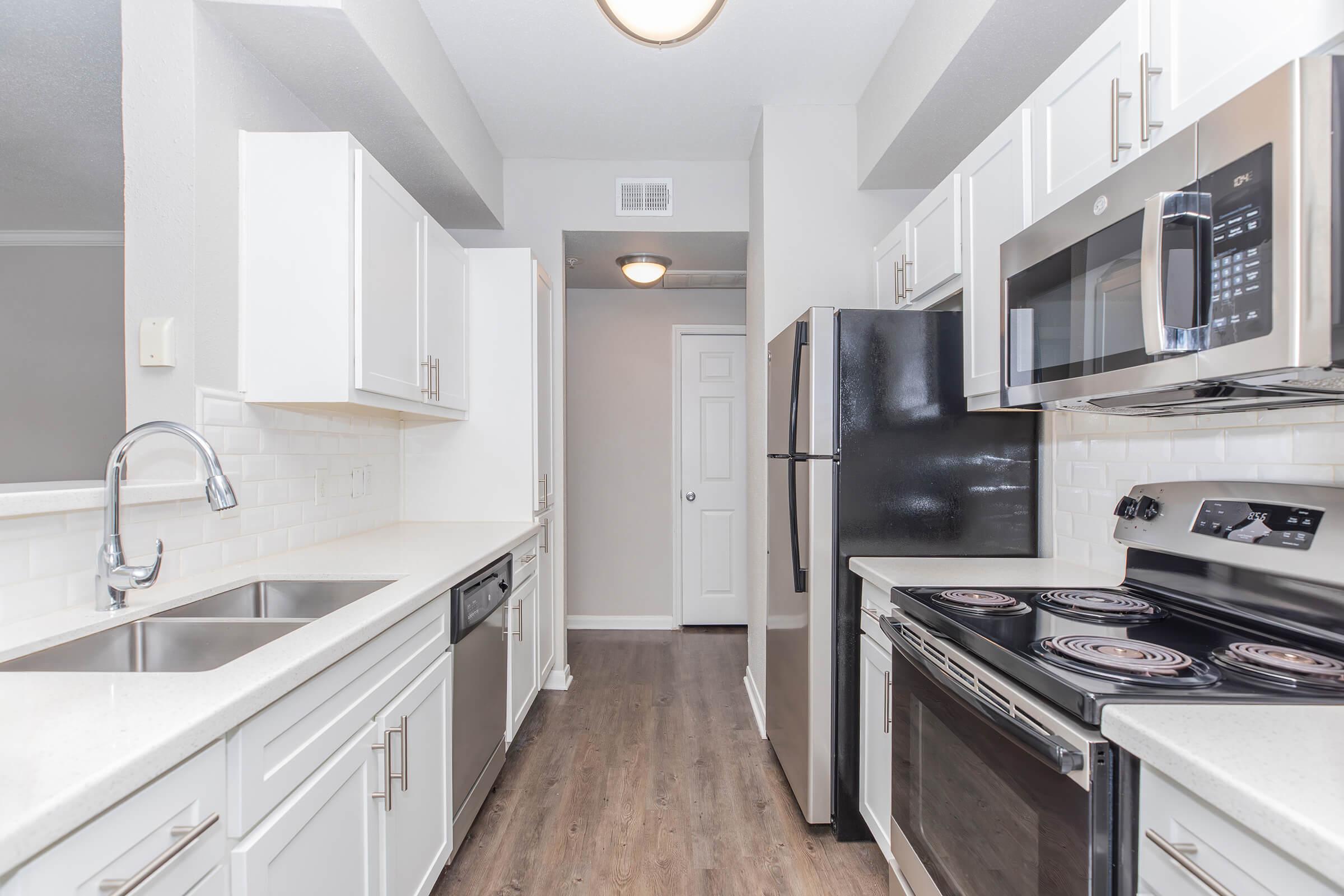 A modern kitchen featuring white cabinets, stainless steel appliances, a double sink, and a dark refrigerator. The countertops are light-colored, and the flooring is a wood-like laminate. The kitchen includes a built-in microwave and an electric stove with four burners. There is a hallway leading to another room.