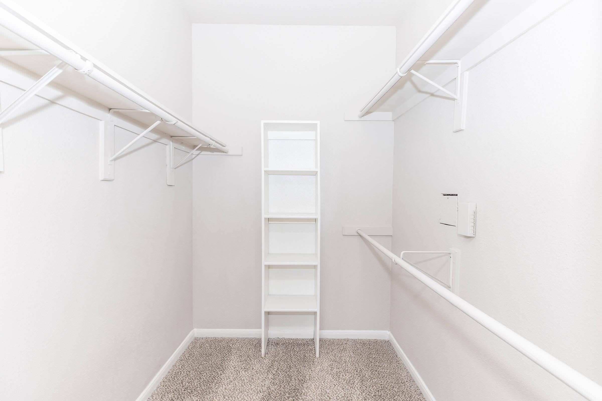 A spacious, empty closet featuring white wall-mounted shelves and a central white shelving unit. The walls are plain and light-colored, while the floor is covered with beige carpet. The overall atmosphere is clean and minimalistic, highlighting the available storage space.