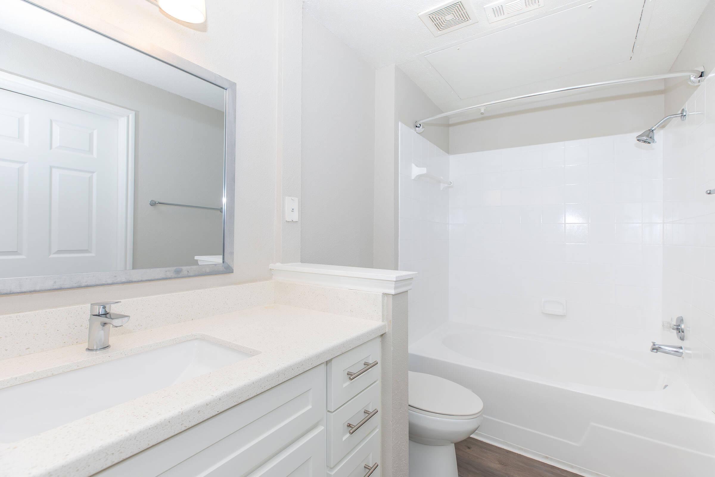 A clean and modern bathroom featuring a white bathtub with a shower, a white vanity with a sink, and a large mirror. The walls are painted in light gray, and there is a door visible leading to another room. The floor has a wood-like finish, adding warmth to the space.
