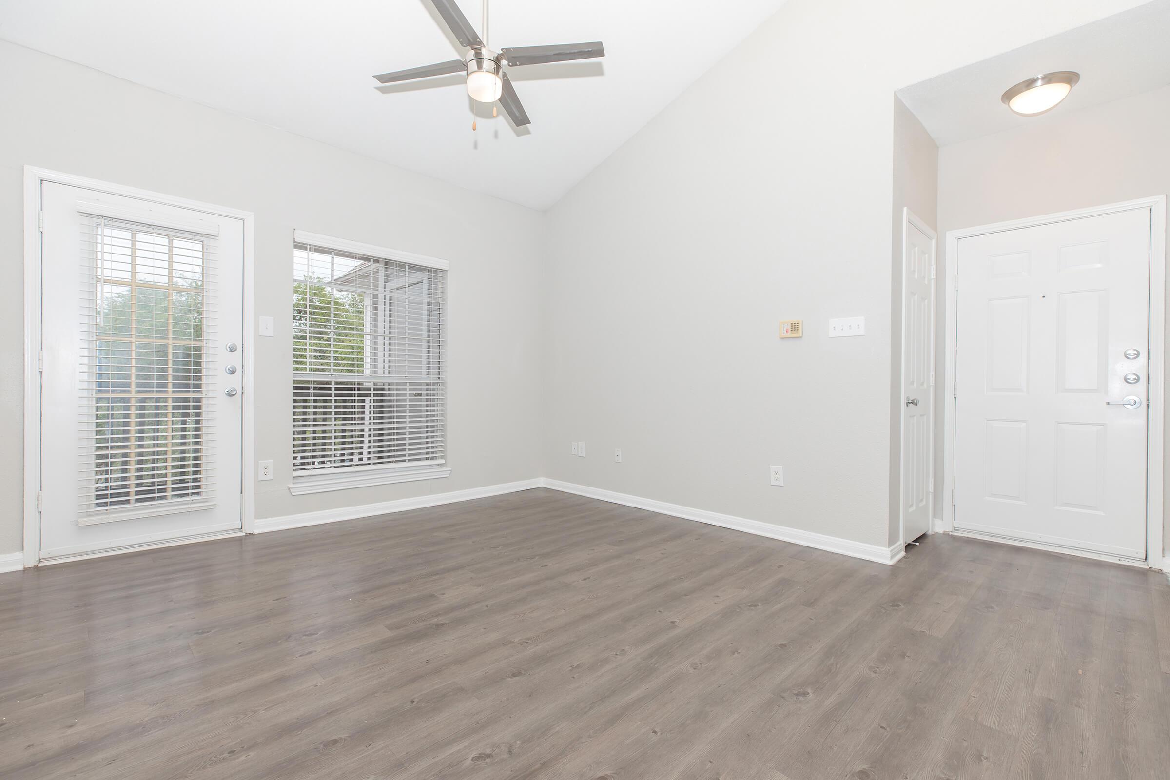 Spacious living area with light gray walls and wood-style flooring. Features a ceiling fan and large windows with blinds allowing natural light. A doorway on the right leads to the entrance, while the left side has sliding glass doors opening to a patio or balcony. The overall atmosphere is bright and inviting.