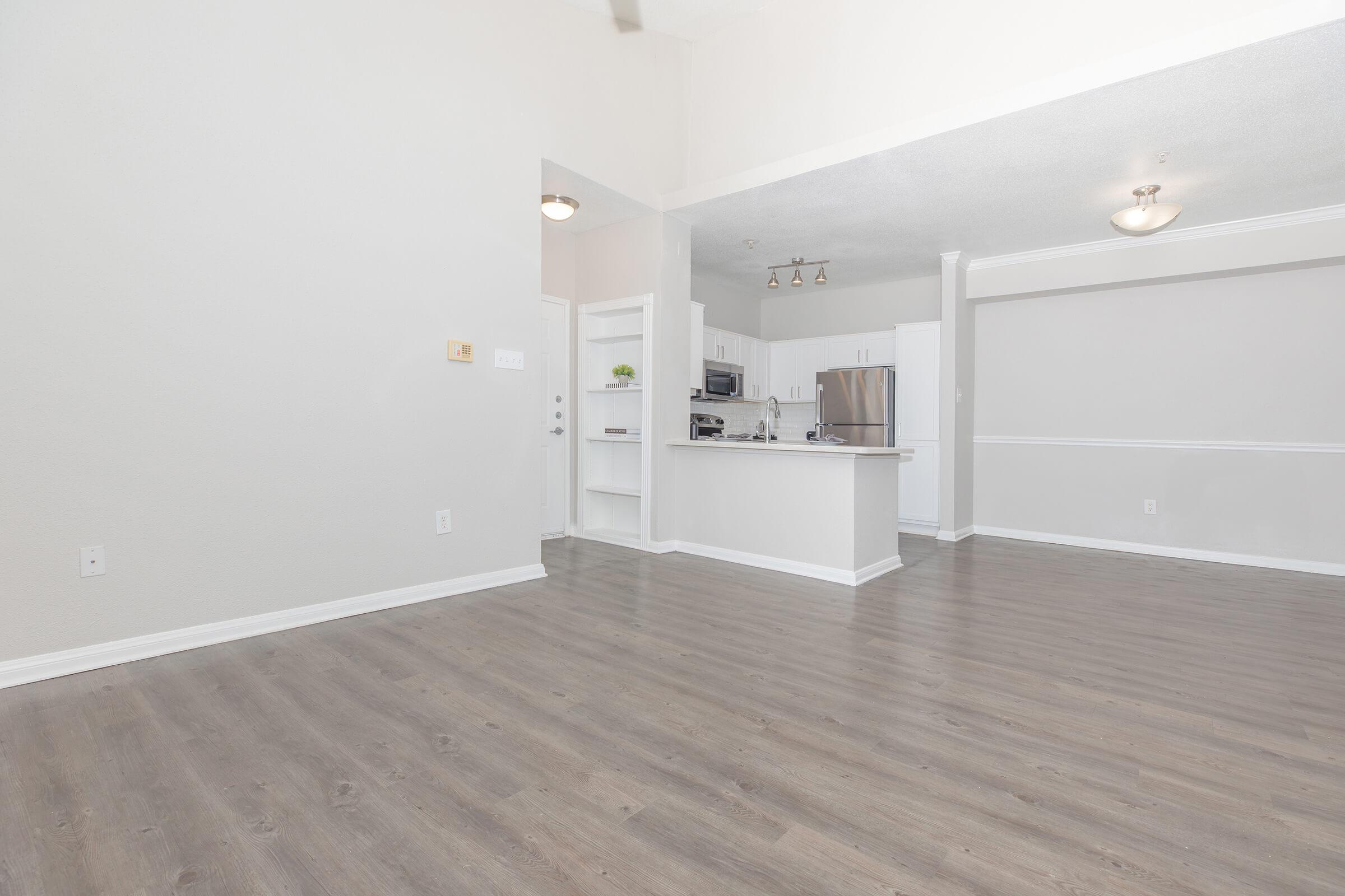 A spacious living area with light-colored walls and wood-like flooring. The kitchen is partially visible, featuring modern appliances and a countertop. The room is well-lit with natural light and has a minimalistic design, creating an open and inviting atmosphere.
