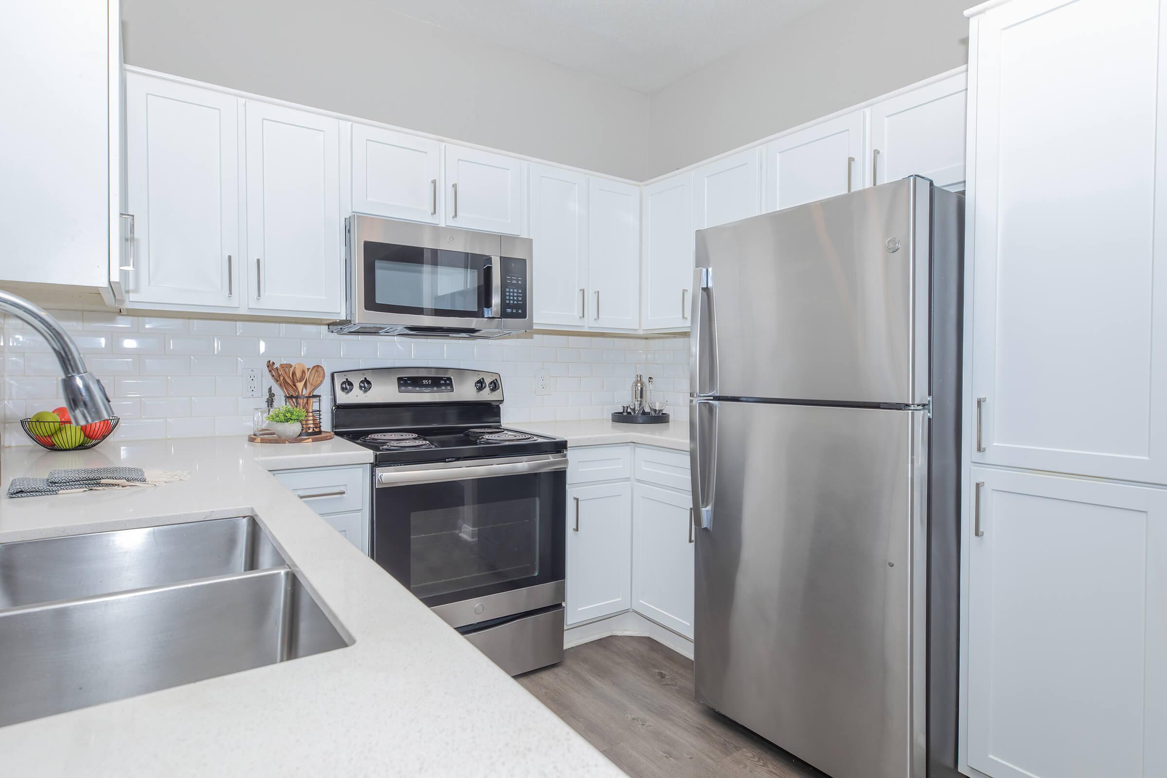 A modern kitchen featuring white cabinets, stainless steel appliances including a microwave, oven, and refrigerator, a double sink, and a countertop. The decor is clean and contemporary, with subtle colors and a minimalistic design.