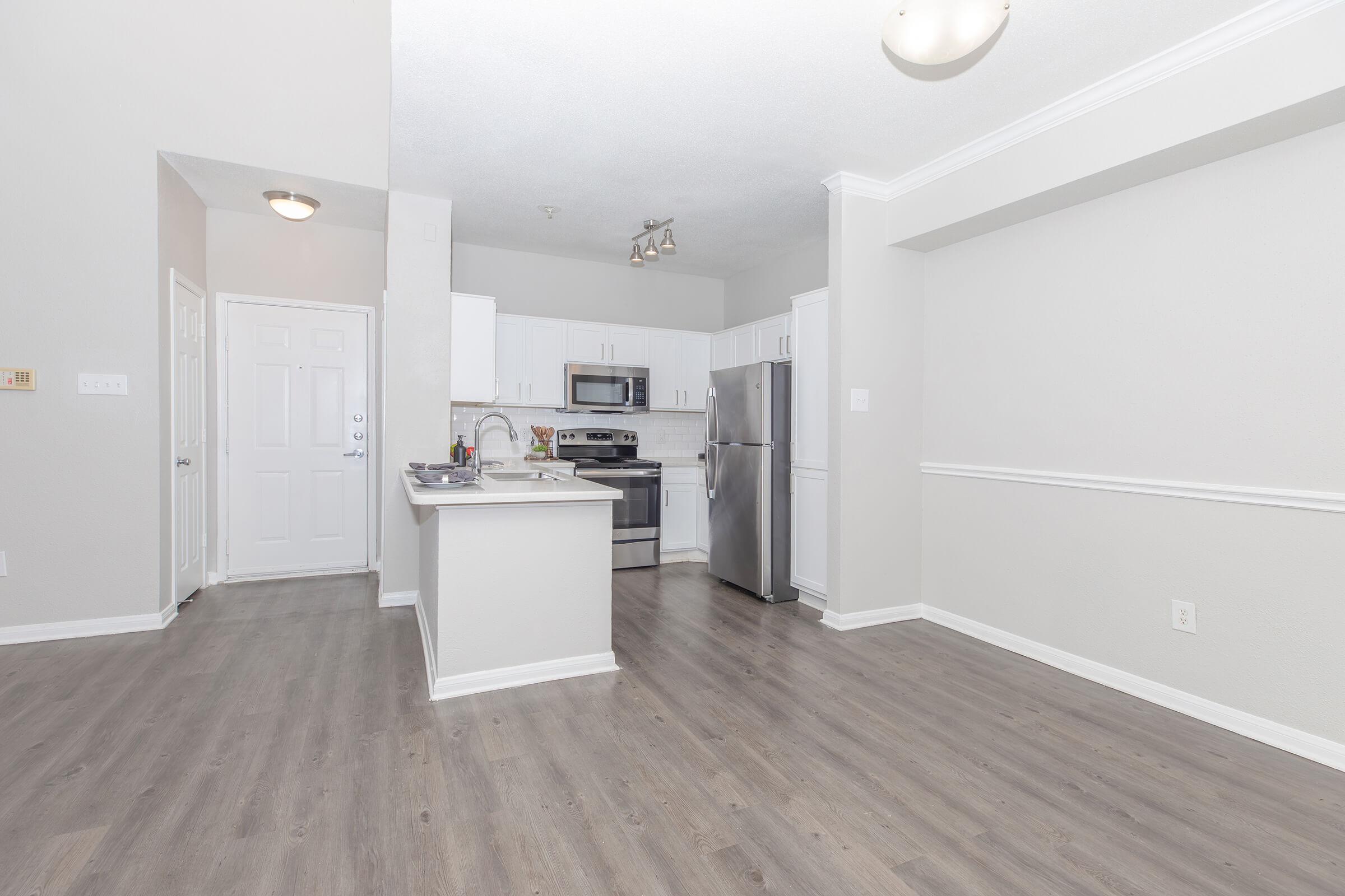 A spacious and modern kitchen and living area featuring light-colored walls, wood-style flooring, stainless steel appliances, and a large window. The kitchen includes a sink, stove, and refrigerator, with an open layout leading to the living space. Bright lighting enhances the overall airy feel.