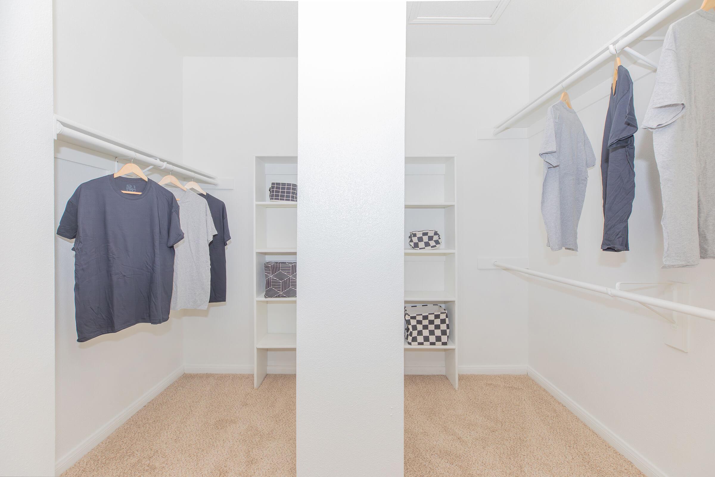 A spacious walk-in closet featuring two sections separated by a white partition. One side has shirts hanging on a rack, while the other side displays neatly arranged shelves with folded items. The walls are white, and the carpet is light beige, creating a clean and organized look.