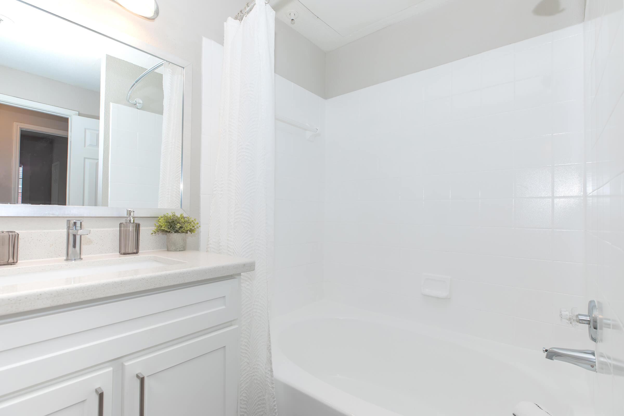 A bright and modern bathroom featuring a white bathtub, sink with a countertop, and a large mirror. The walls and tiles are white, creating a clean and airy atmosphere. A small plant adds a touch of greenery on the counter, and a shower curtain is visible next to the tub.