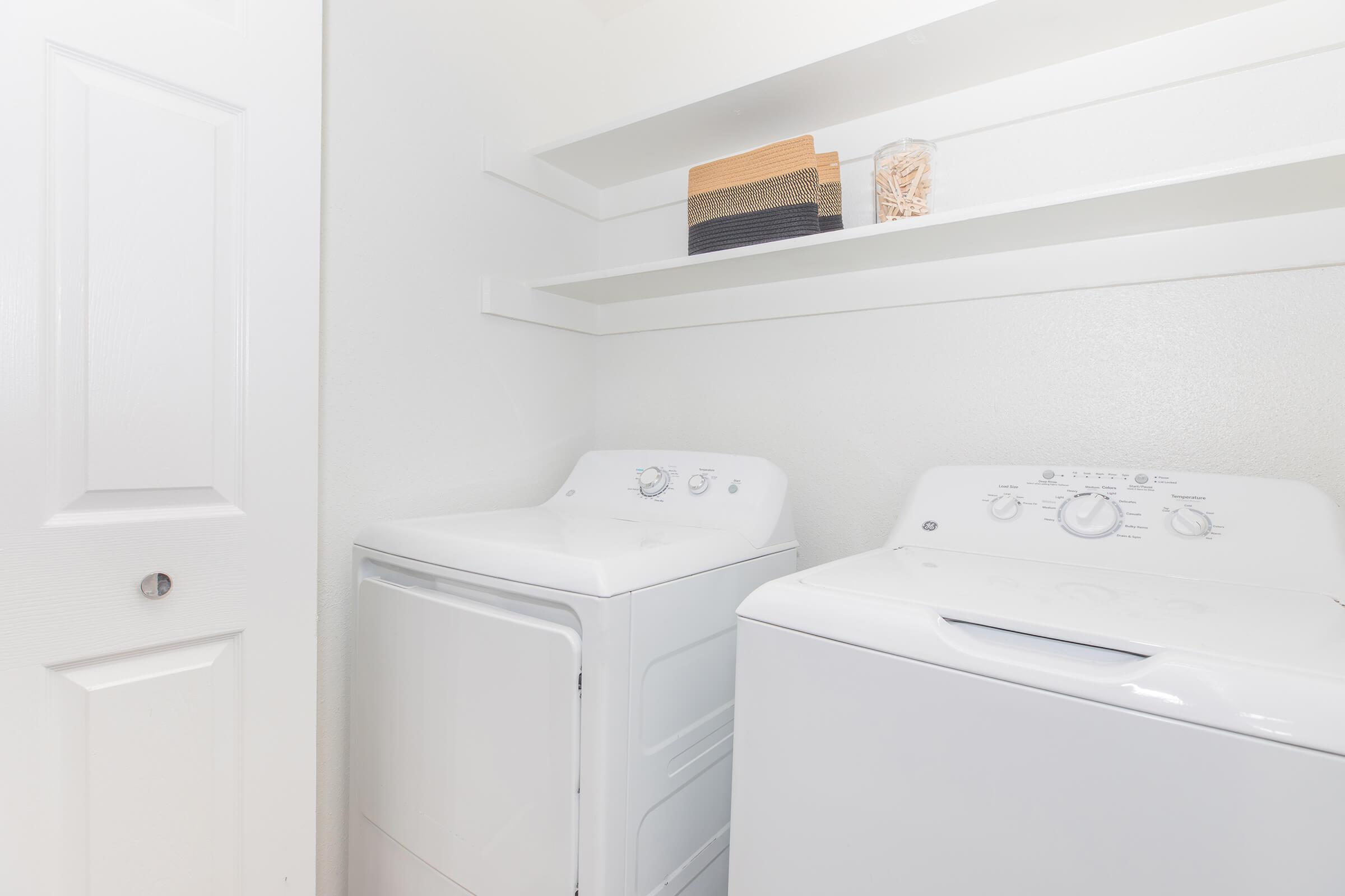 A clean laundry room featuring a white washing machine and dryer side by side, with white walls and minimalist shelving above. The shelves hold a few storage boxes and clothespins. A closed door is visible, adding to the organized and tidy appearance of the space.