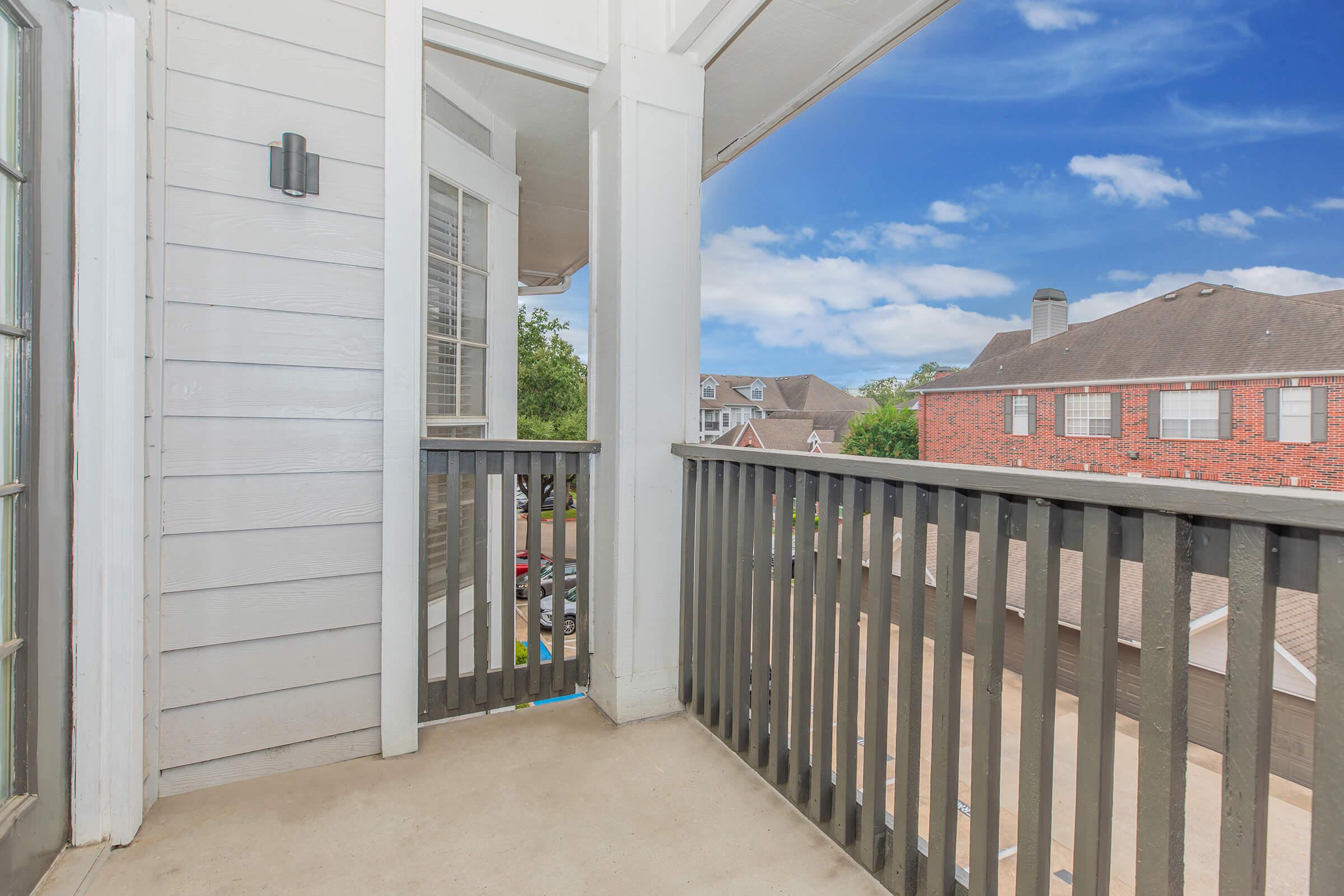 A balcony view showcasing a railing and wall with light-colored siding. The sky is bright blue with some clouds, while a few buildings are visible in the background, featuring red brick and gray exteriors. The area appears well-maintained and inviting, ideal for outdoor relaxation.