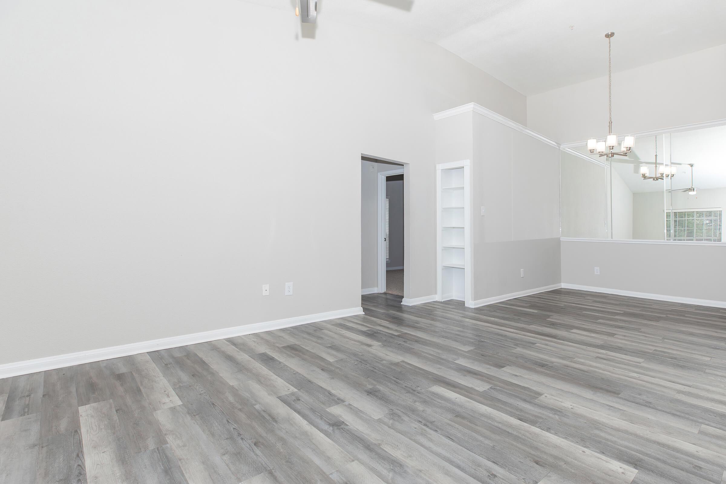 Bright, spacious interior of a modern living room featuring light gray walls and a hardwood floor. A doorway leads to another room, while a set of shelves and a chandelier adds style. Large windows reflect natural light, enhancing the open feel of the space.