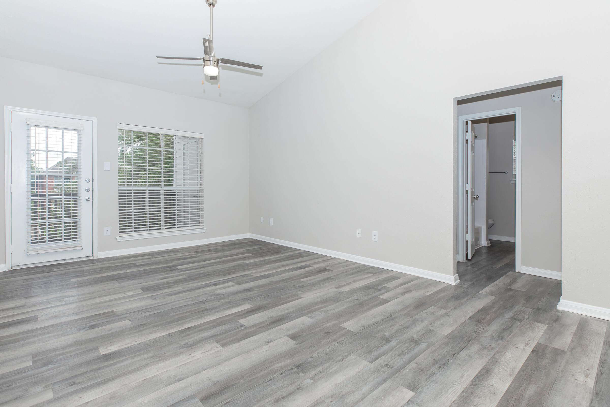 An empty room with light gray walls and a high ceiling. The floor features gray laminate planks. A ceiling fan is mounted in the center, and there are large windows with white blinds on one wall. A doorway leads to another space on the right, creating an open and spacious feel.