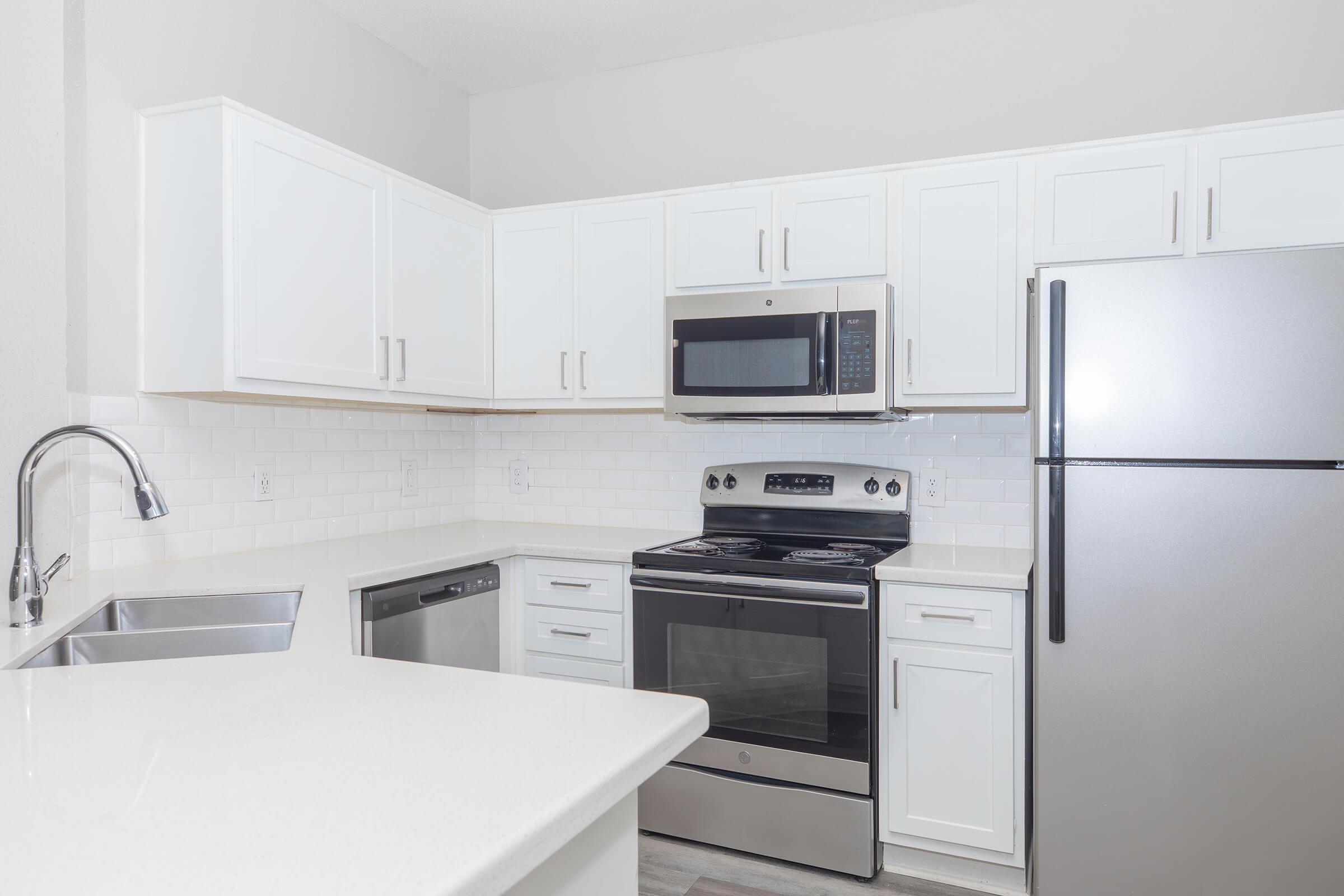 A modern kitchen featuring white cabinetry, a stainless steel refrigerator, a microwave above a black stove, a silver dishwasher, and a sink with a sleek faucet. The countertops are light-colored, and the backsplash consists of white tiles, creating a bright and clean aesthetic.