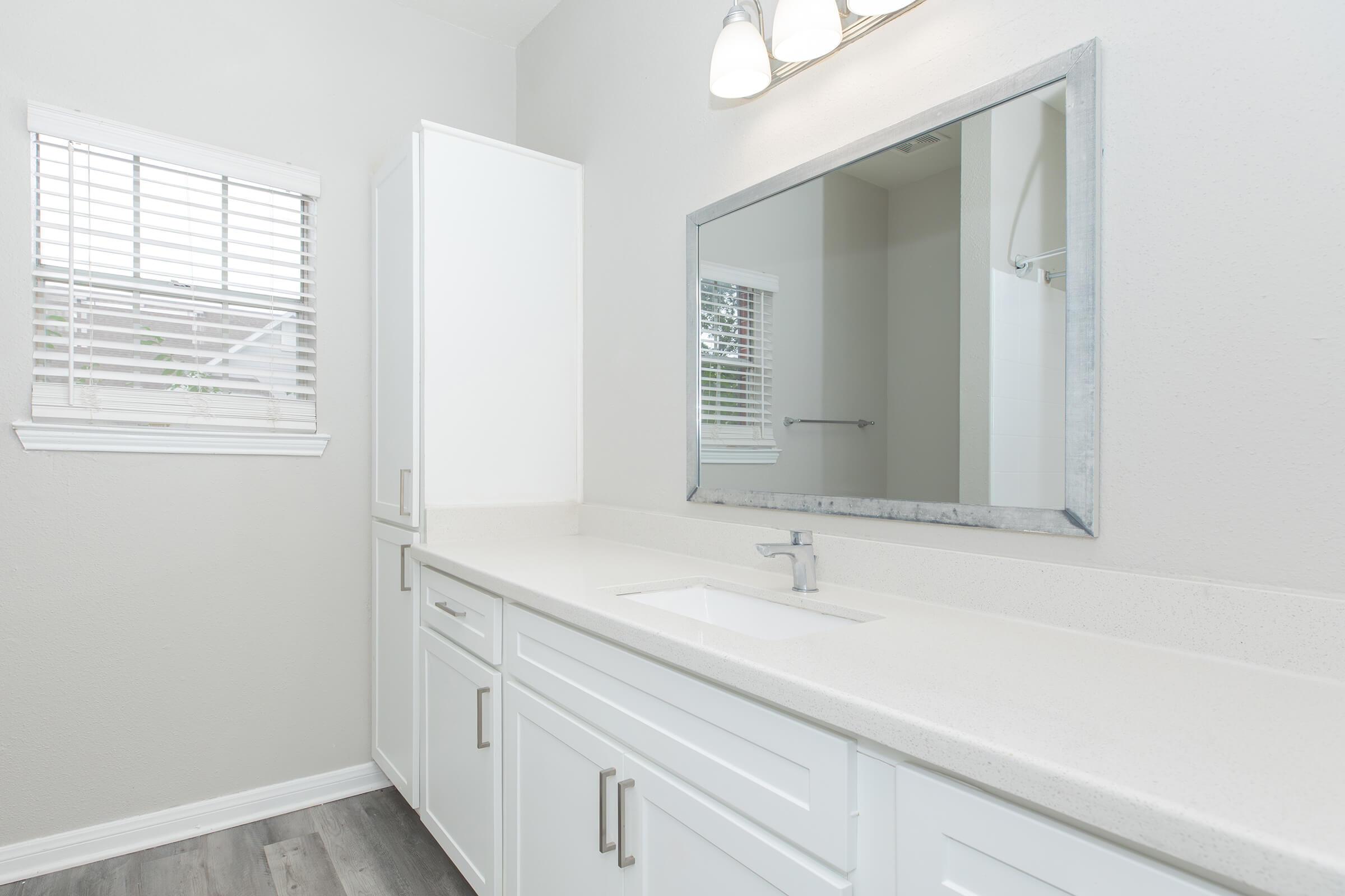 A modern bathroom featuring a sleek white countertop, a large rectangular mirror with a metal frame, and white cabinetry. Natural light filters through a window with blinds, illuminating the space. The walls are painted a soft neutral color, enhancing the bright and clean aesthetic.