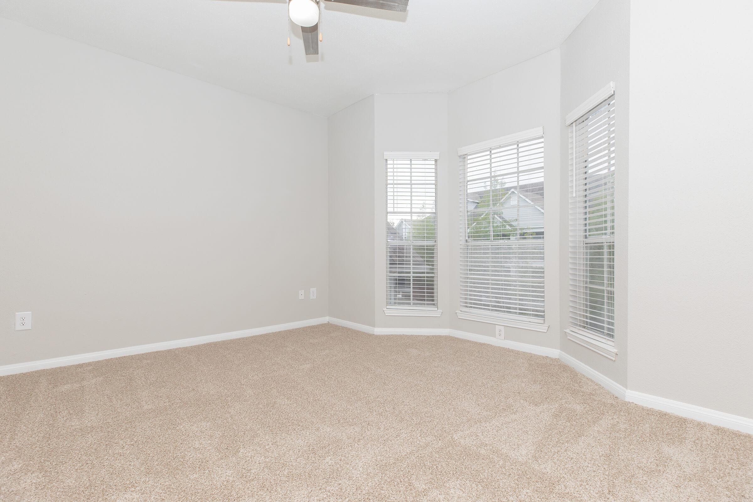 Empty room with beige carpet, light gray walls, and a ceiling fan. The room features large windows with white blinds, allowing natural light in. One corner has a bay window area. Overall, the space is bright and inviting, suitable for various furnishings and decor.