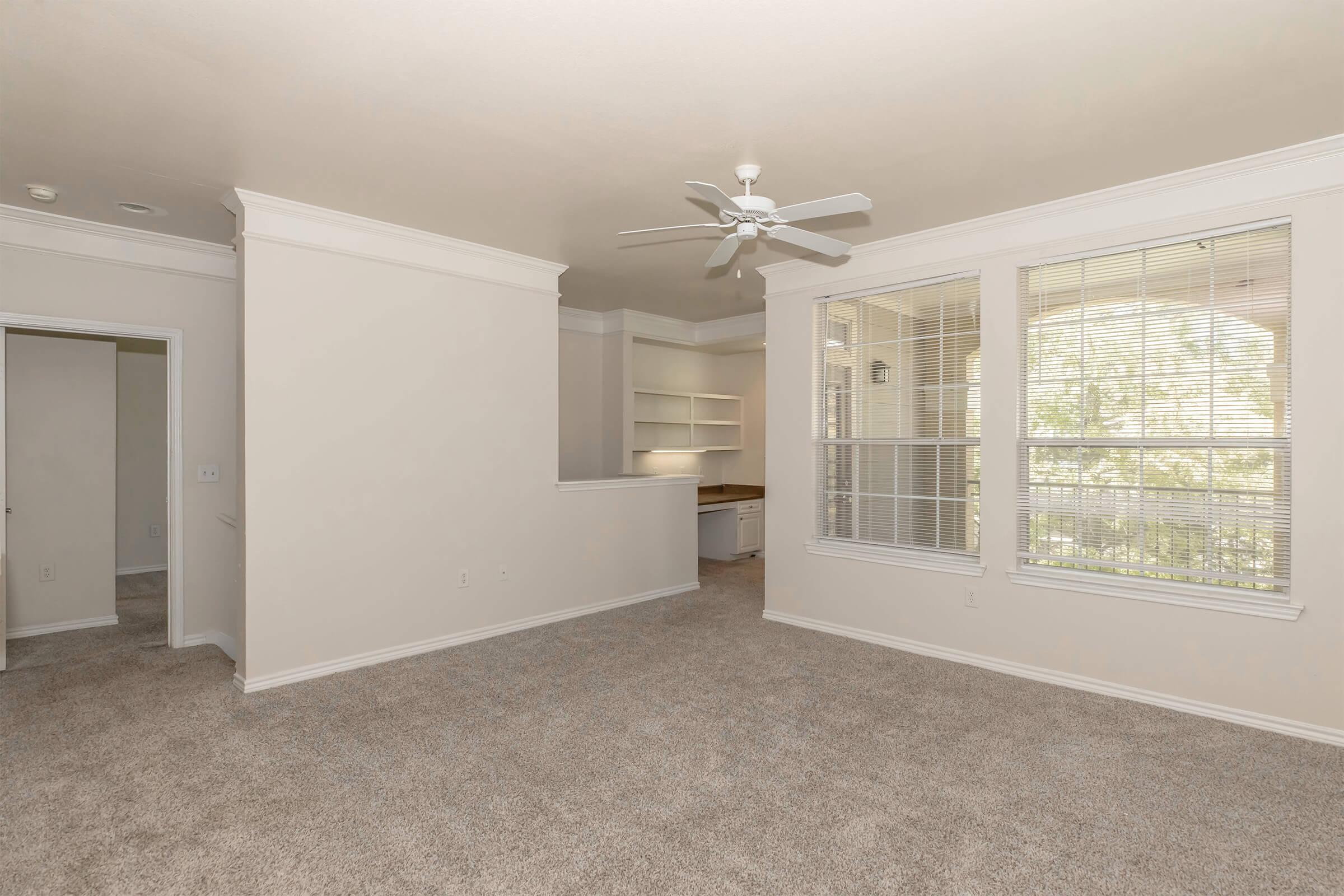 A well-lit, spacious living area featuring light-colored walls, a ceiling fan, and large windows with blinds. The floor is covered in beige carpet, and there is an open space leading to a small kitchen area with built-in shelves. A door on the left indicates entry to another room.
