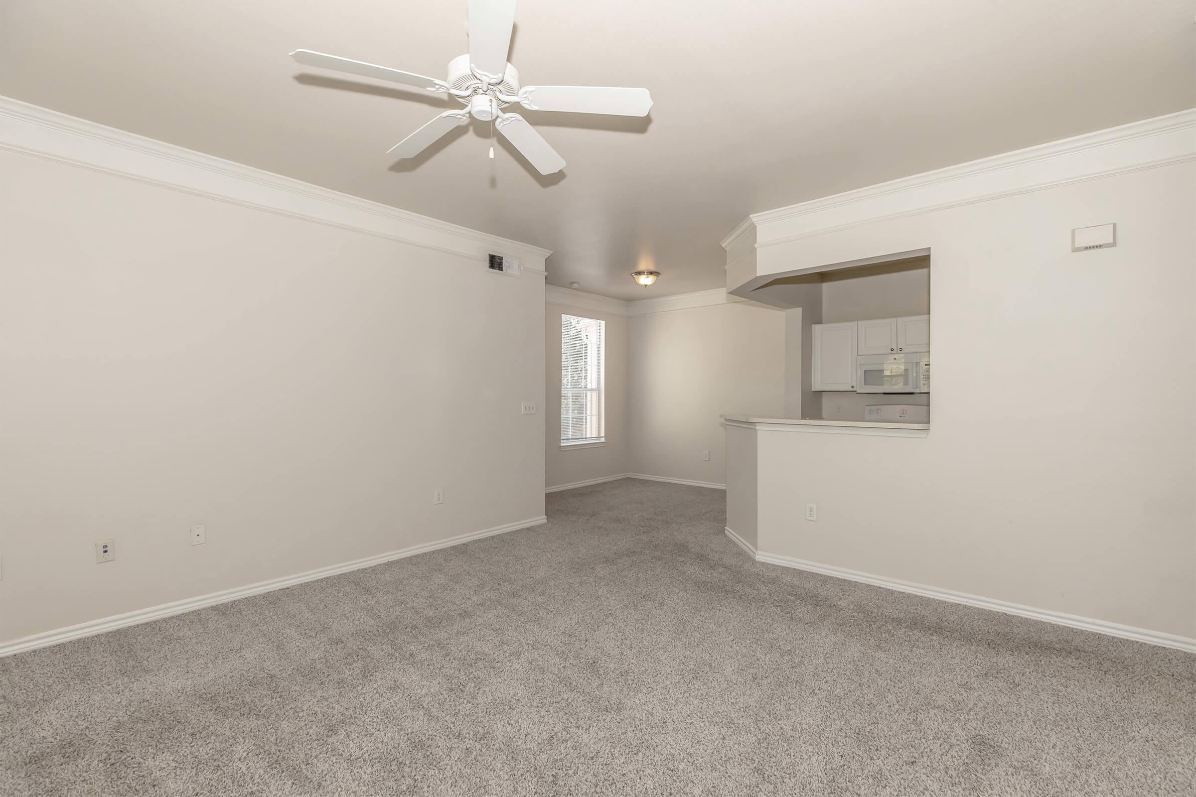 A spacious and empty living area with light-colored walls and plush carpet. A ceiling fan is visible, and there's a small adjacent kitchen area with white cabinetry. Natural light filters through a window, illuminating the room's neutral tones.
