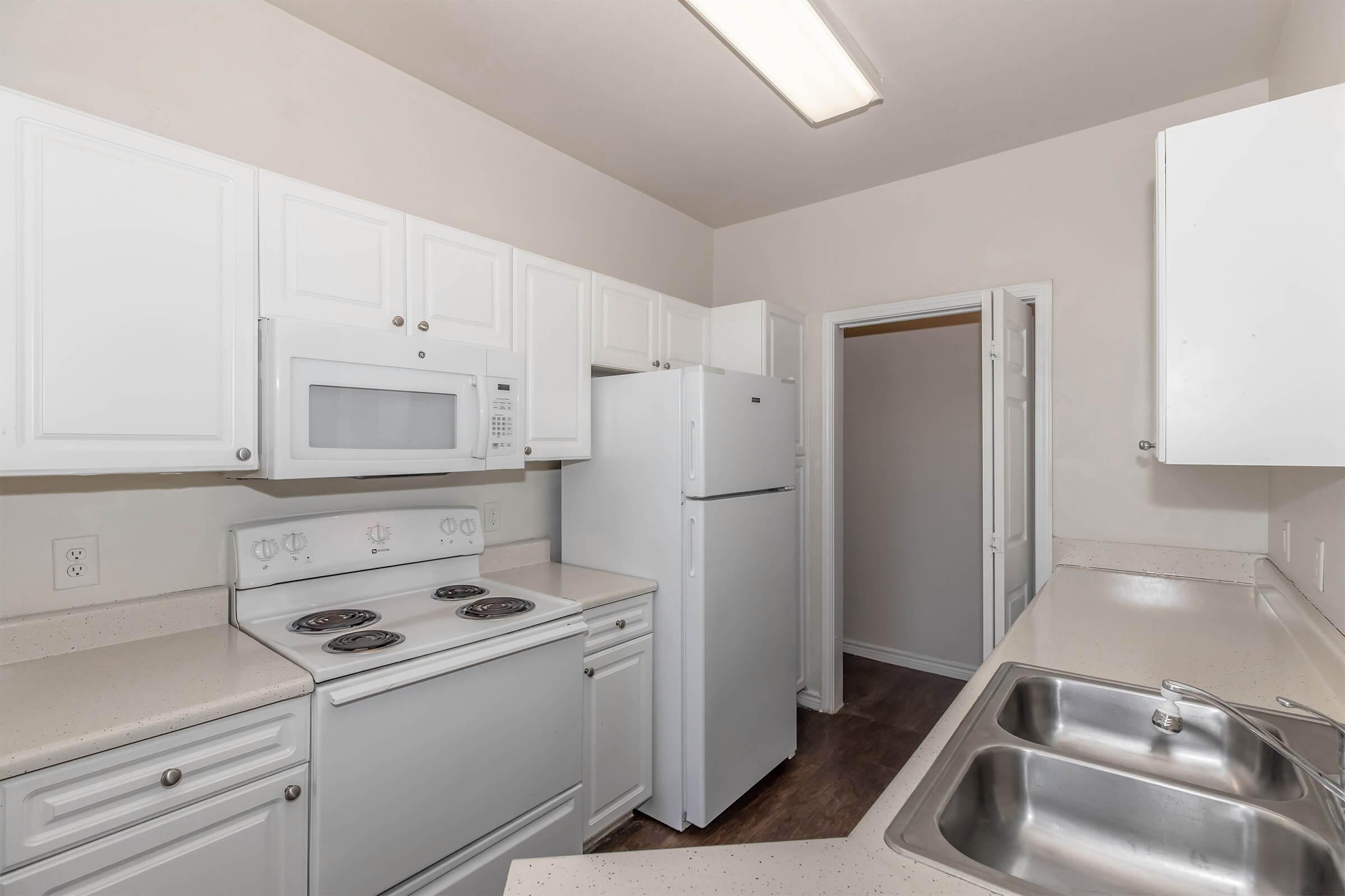 A modern kitchen with white cabinetry, featuring a microwave, a refrigerator, and a stove with four burners. The countertops are light-colored, and there's a double sink visible. A door leads to a storage area or pantry. The overall design is simple and functional, with a clean and bright atmosphere.
