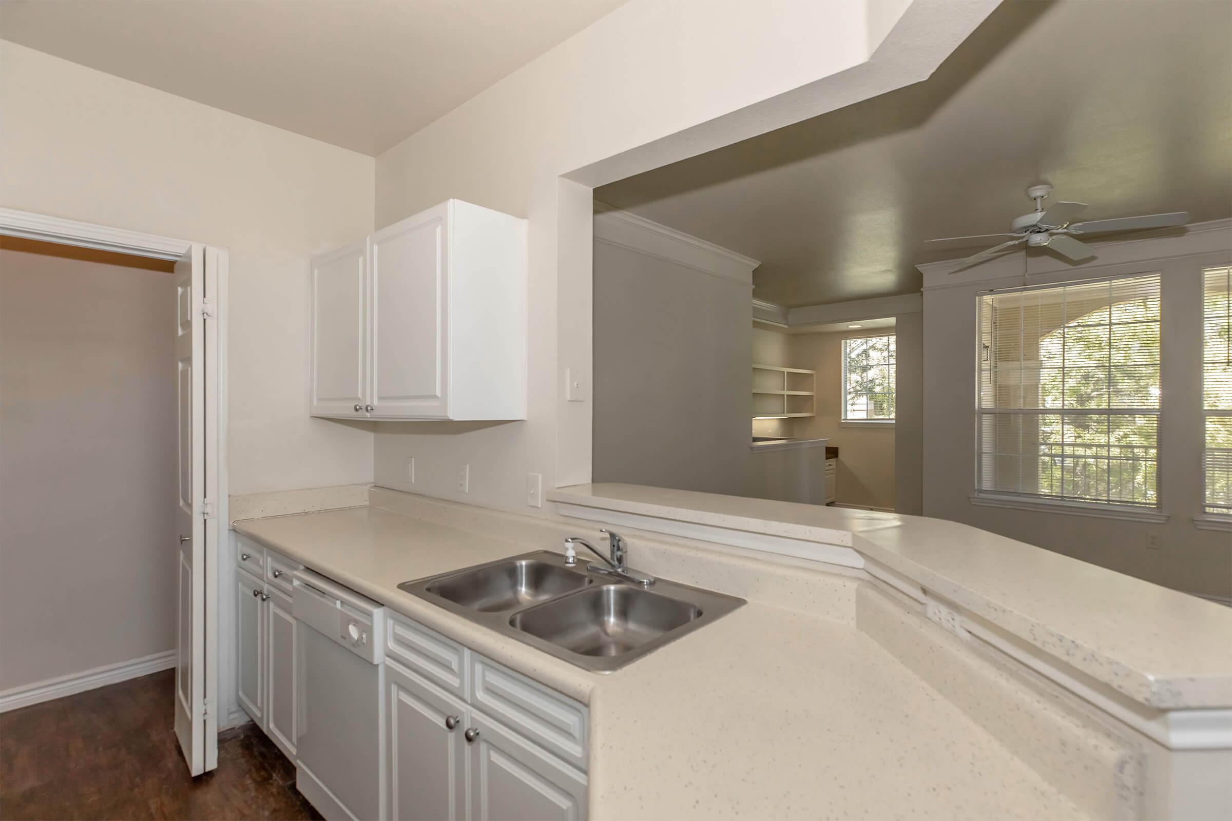 A modern kitchen featuring light-colored cabinets, a double sink, and a spacious countertop. The open layout connects to a living area with large windows allowing natural light, creating a bright and airy atmosphere.